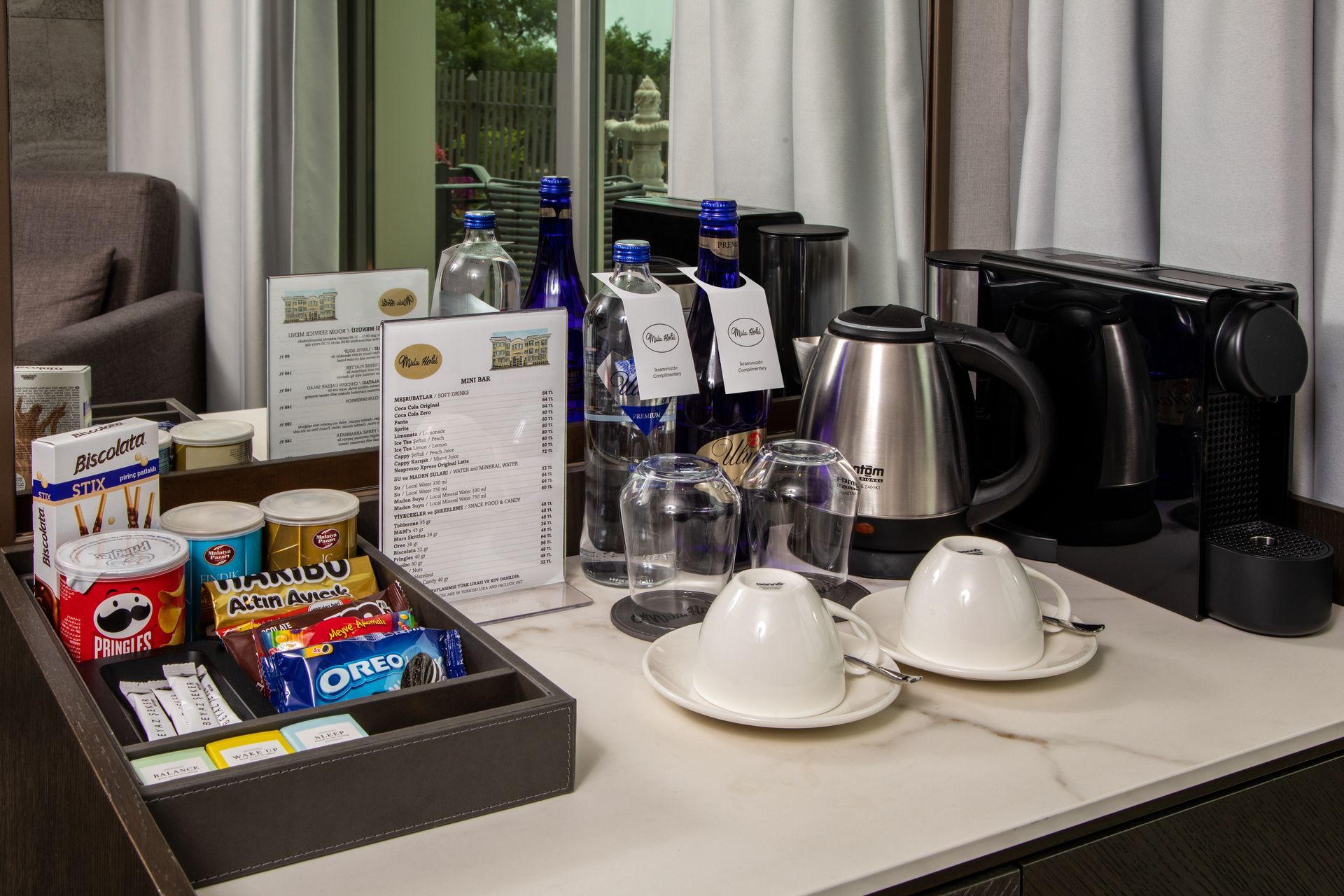 Hotel room coffee and snack station with kettle, cups, bottled water, and packaged treats