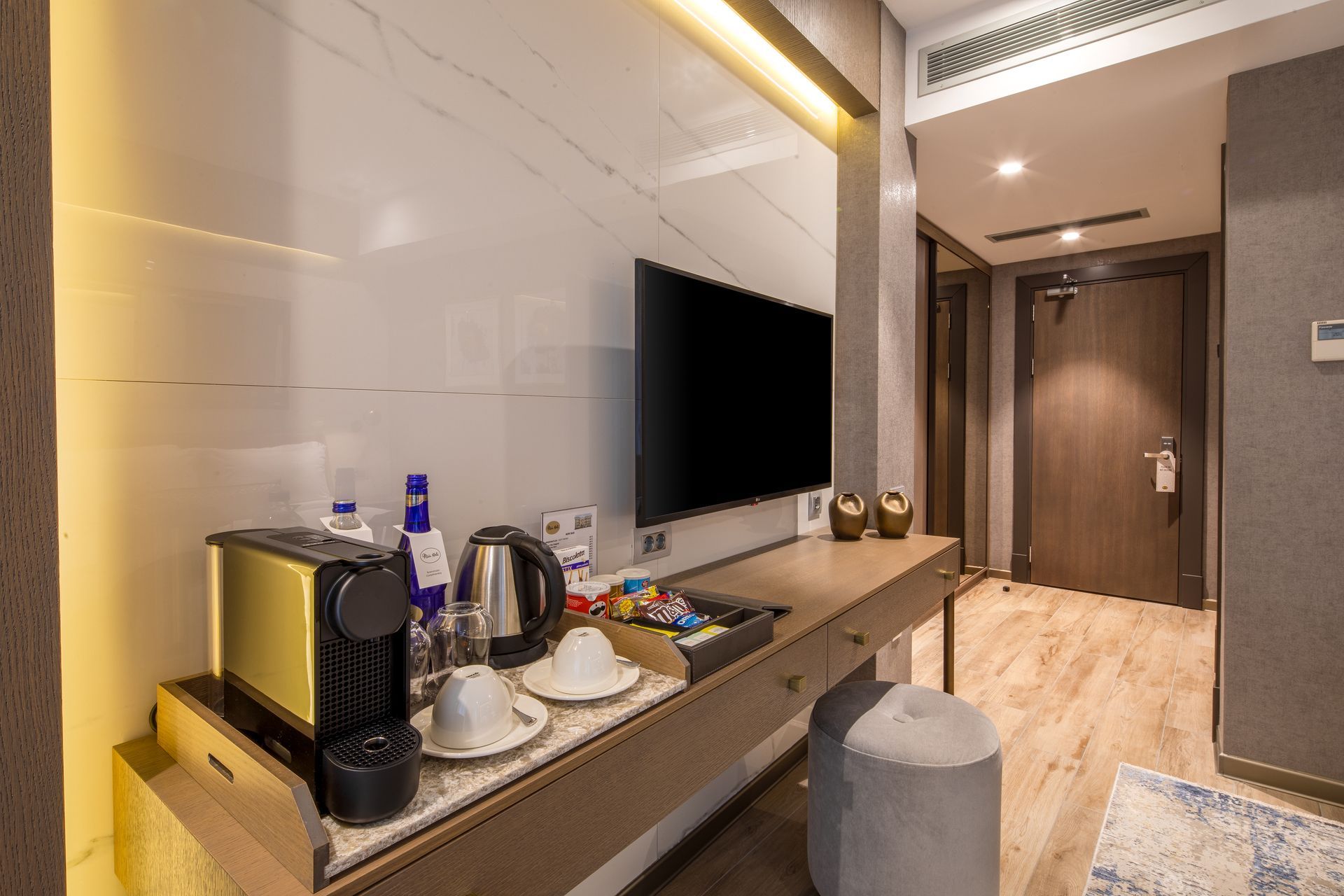 Modern hotel room with TV, minibar tray, and luggage stool beside a wood-paneled hallway