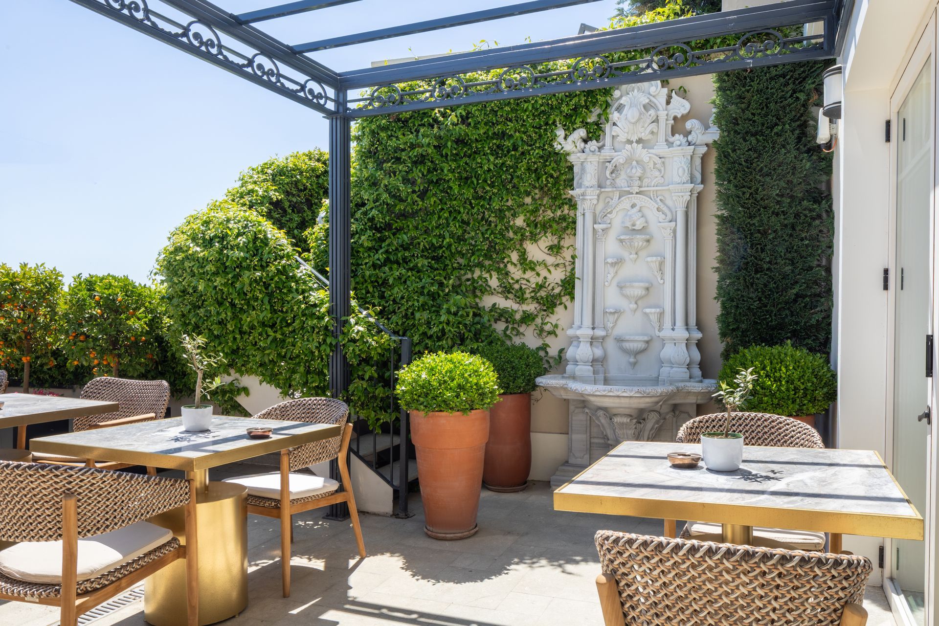 Sunlit outdoor cafe terrace with wicker tables, ivy wall, and white fountain centerpiece