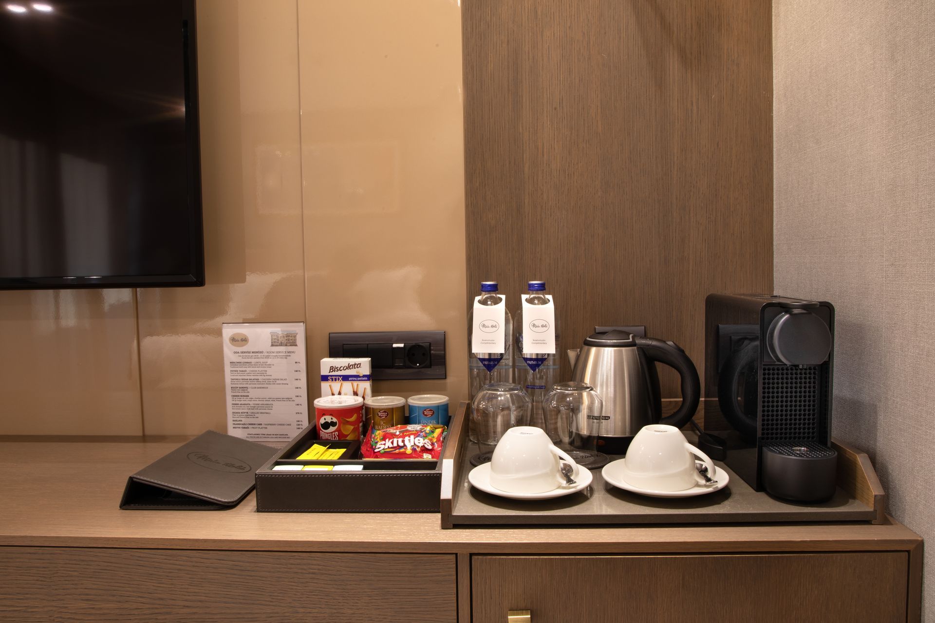 Hotel room beverage station with TV, kettle, cups, bottled water, snacks, and coffee maker on a countertop