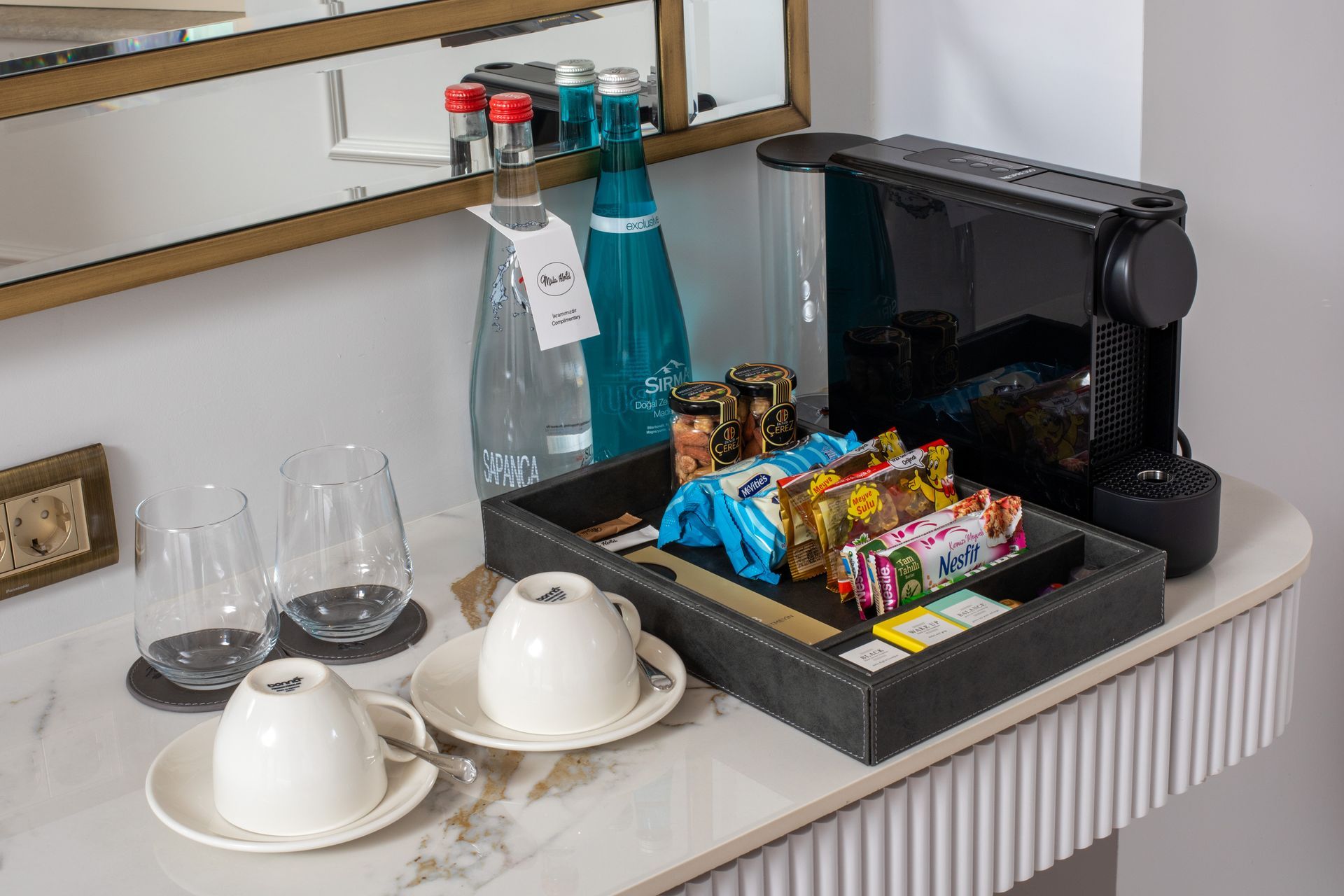 Coffee station with a pod machine, cups, water bottle, and assorted snacks on a tray
