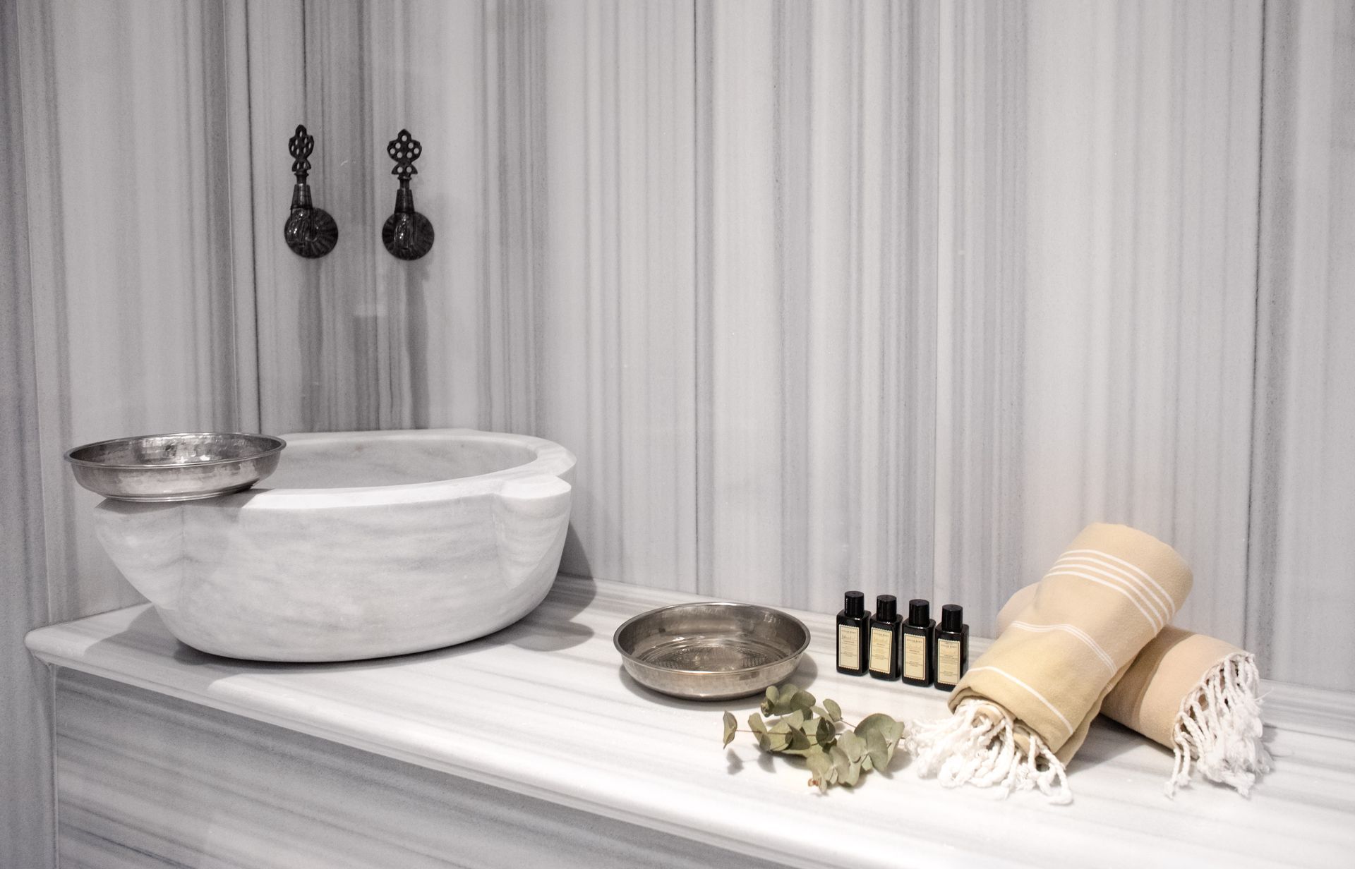 Spa setup with marble bowl, brass tray, essential oil bottles, towels, and greenery on a striped tablecloth
