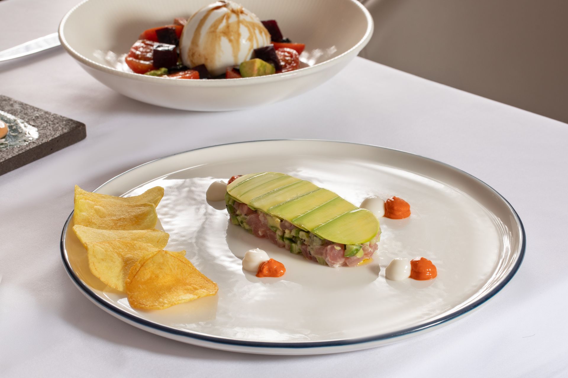 Gourmet plated dish with avocado slices and chips, with a dessert bowl in the background