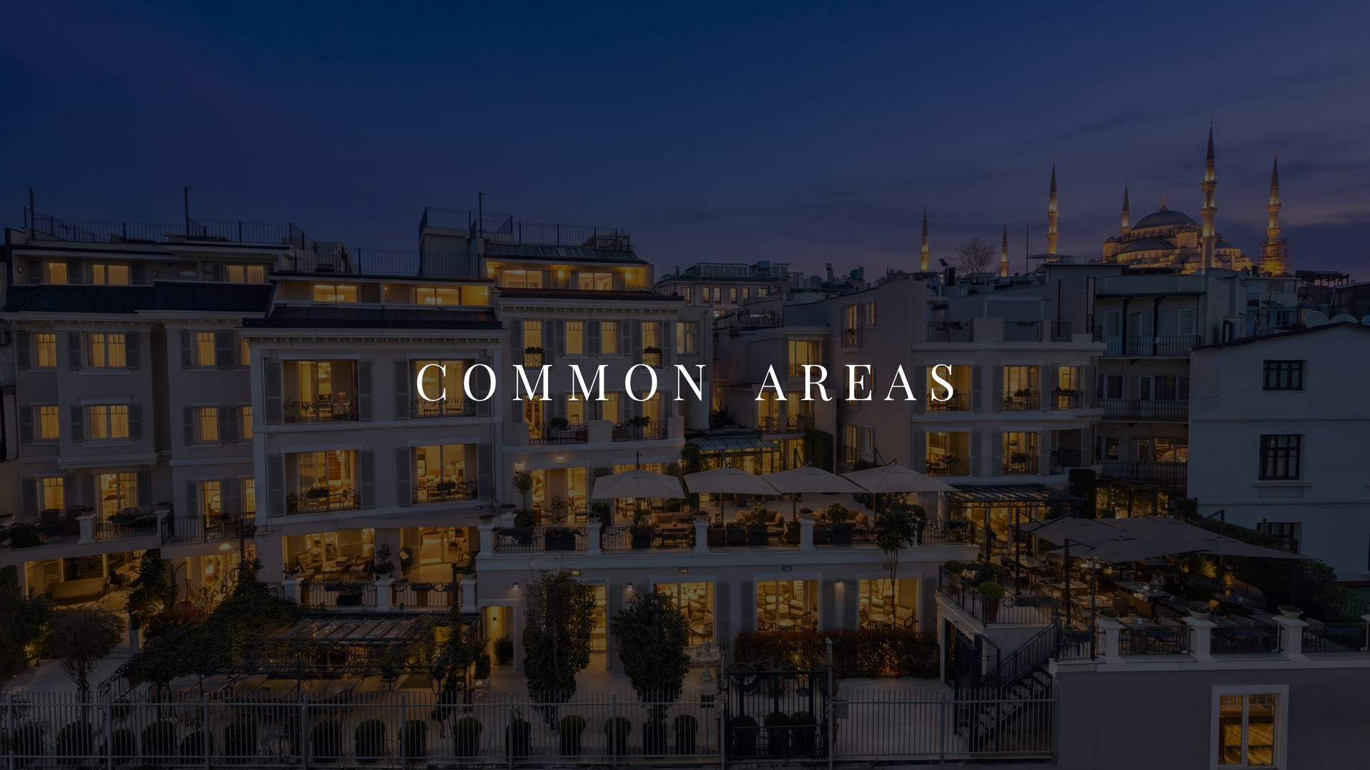 Dusk cityscape of a rooftop terrace and apartment buildings, with lit windows and a distant mosque skyline.