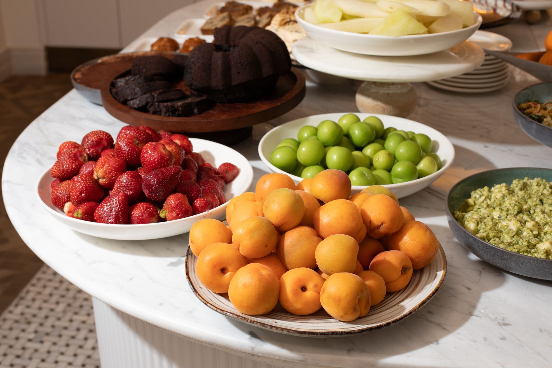 Mula Hotel | A marble table with a spread of various fresh fruits and desserts in bowls.