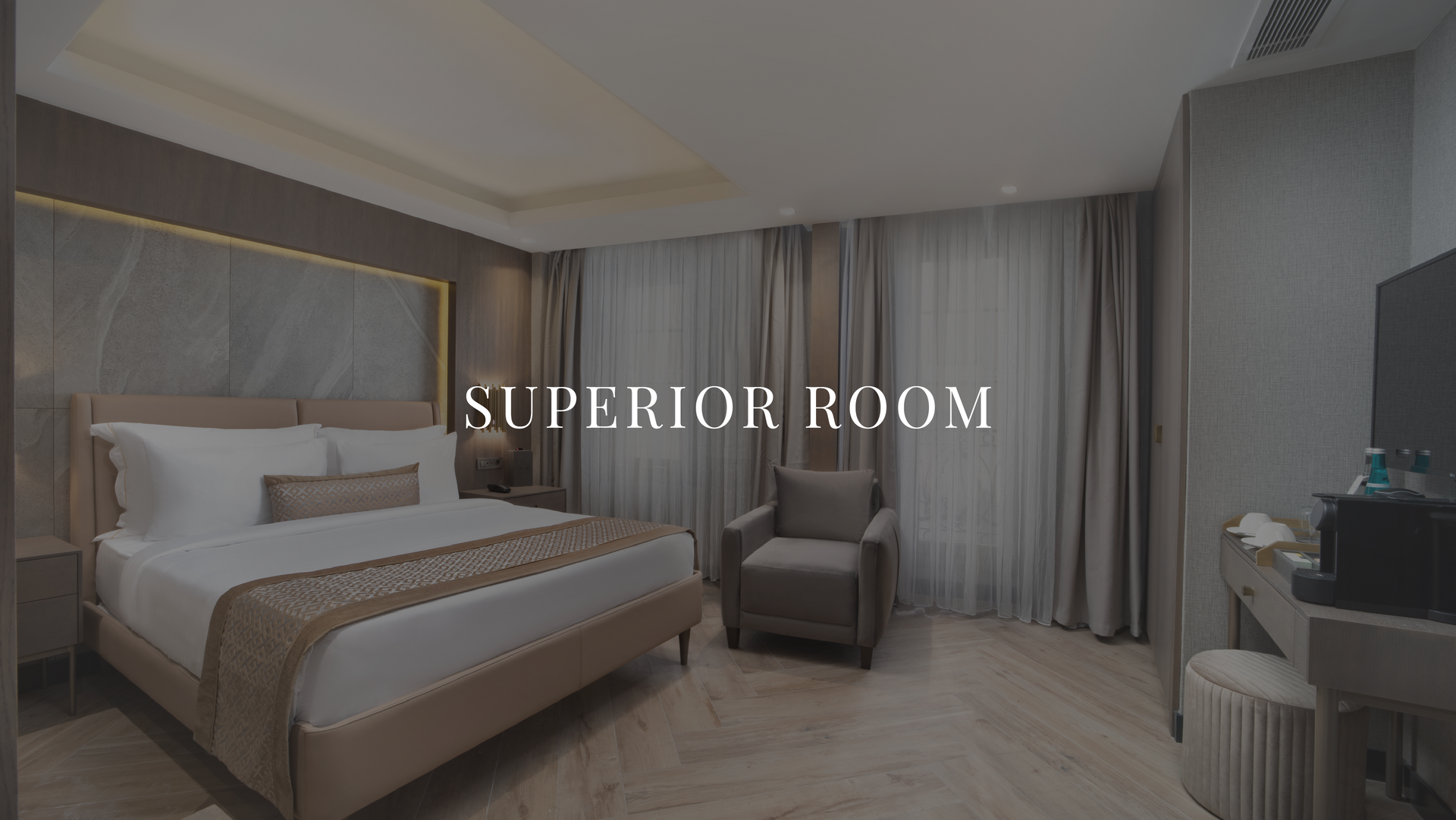 Modern hotel room with king bed, armchair, desk, and gray curtains; “SUPERIOR ROOM” text overlay. | Mula Hotel