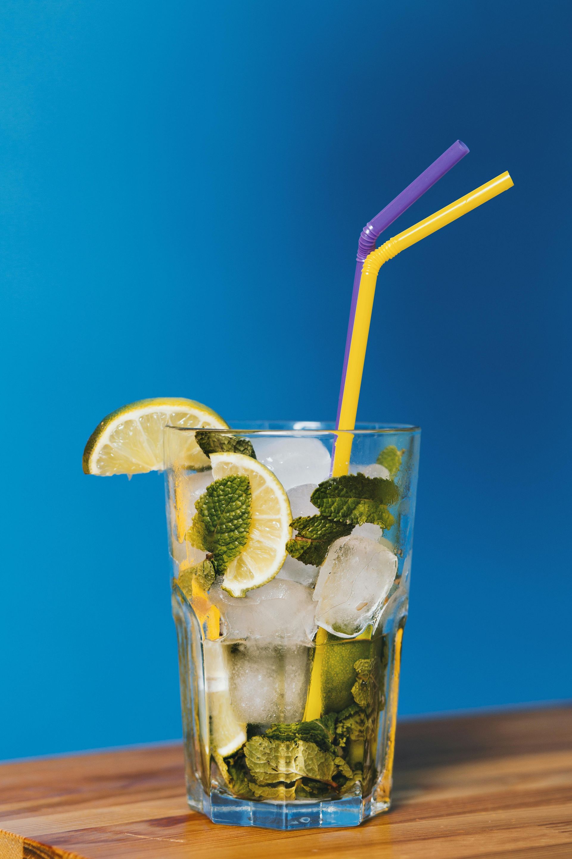 Mula Hotel | Mojito cocktail in a glass with lime, mint, ice, and straws against a blue background.