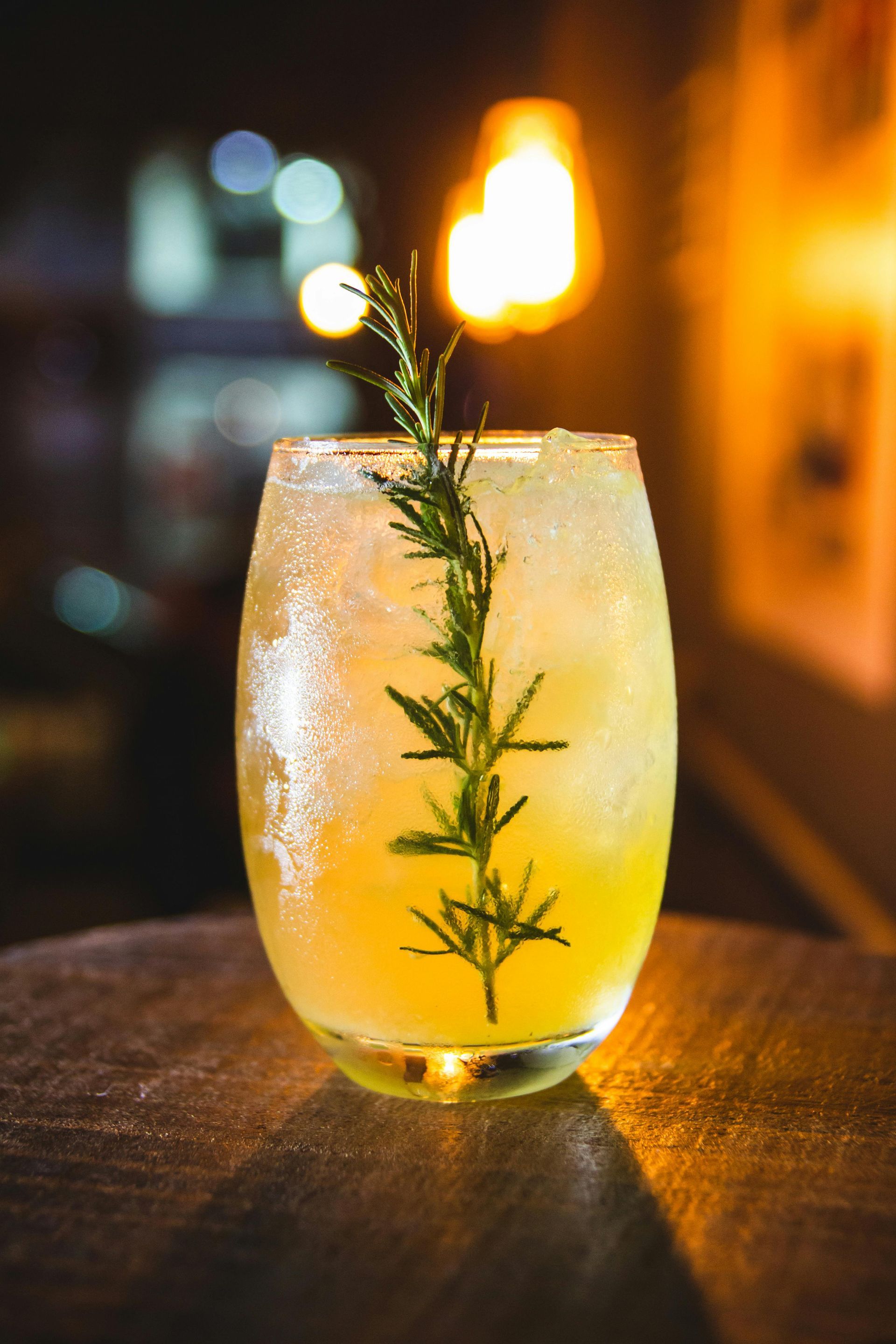 Mula Hotel | Cocktail in a glass with a rosemary sprig on a wooden surface, with blurred city lights in the background.