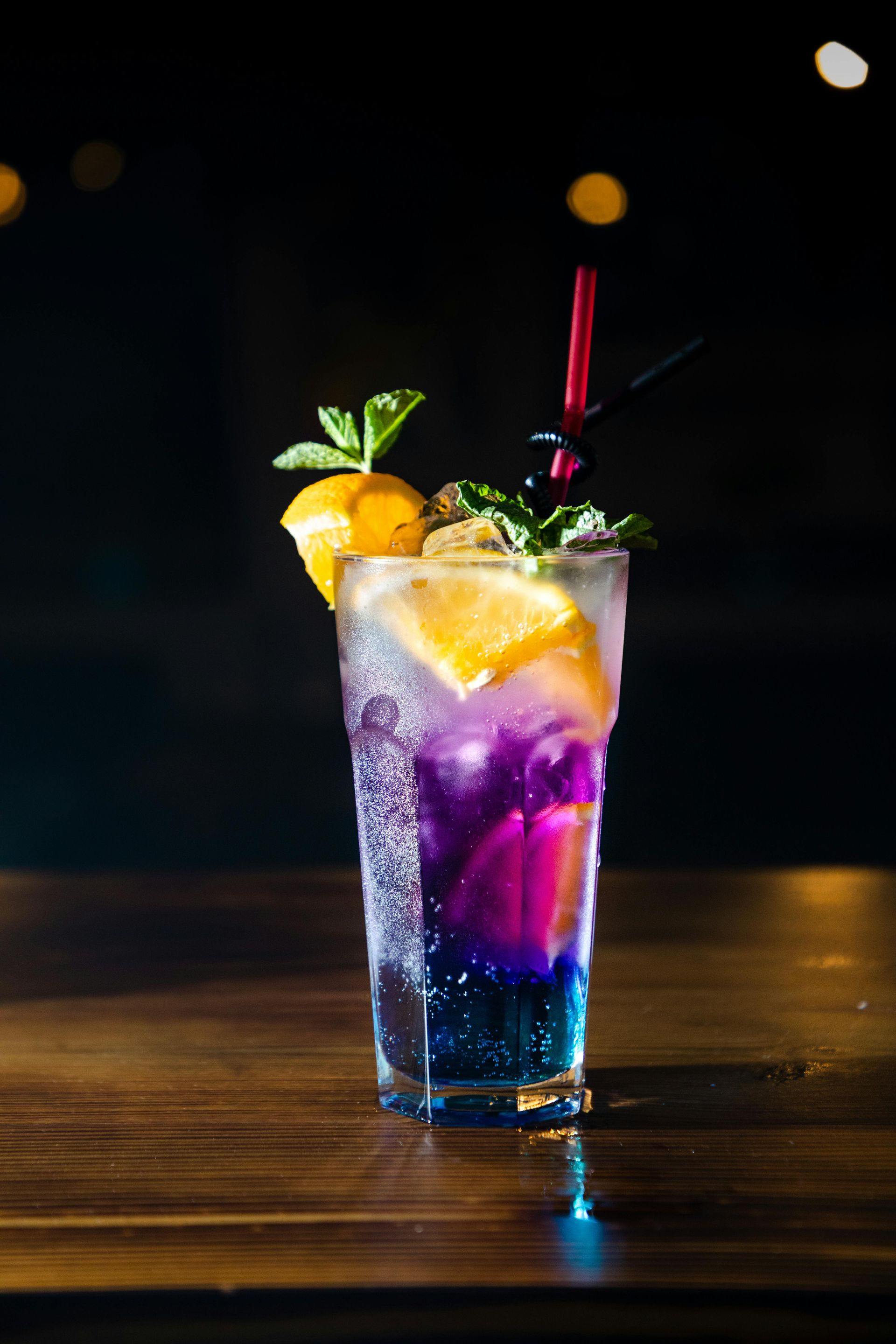 Mula Hotel |Colorful cocktail in a tall glass, garnished with orange slice, mint, and a straw, on a wood surface.
