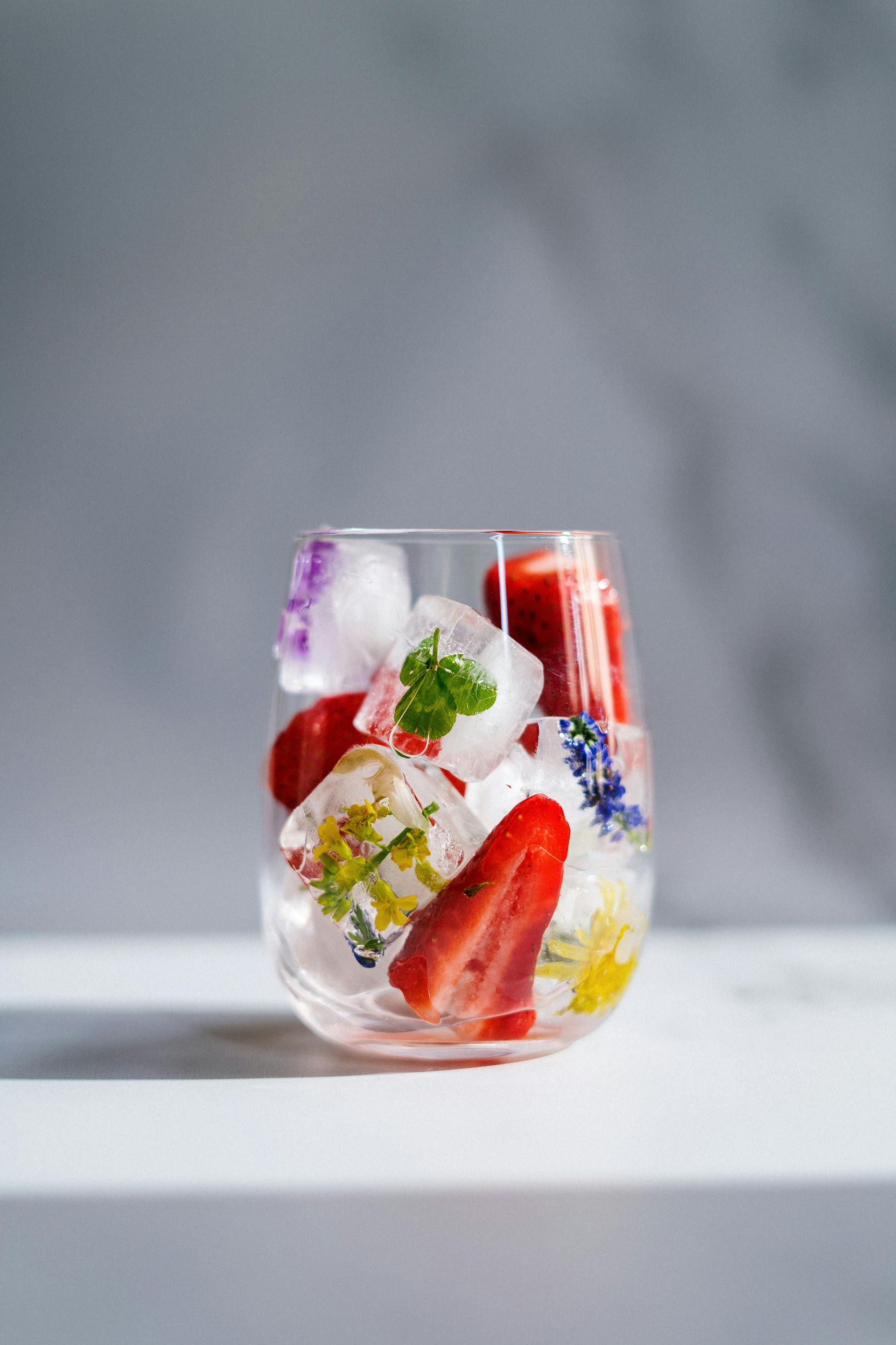 Mula Hotel |Clear glass filled with ice cubes, strawberries, and flowers, on a white surface against a gray background.