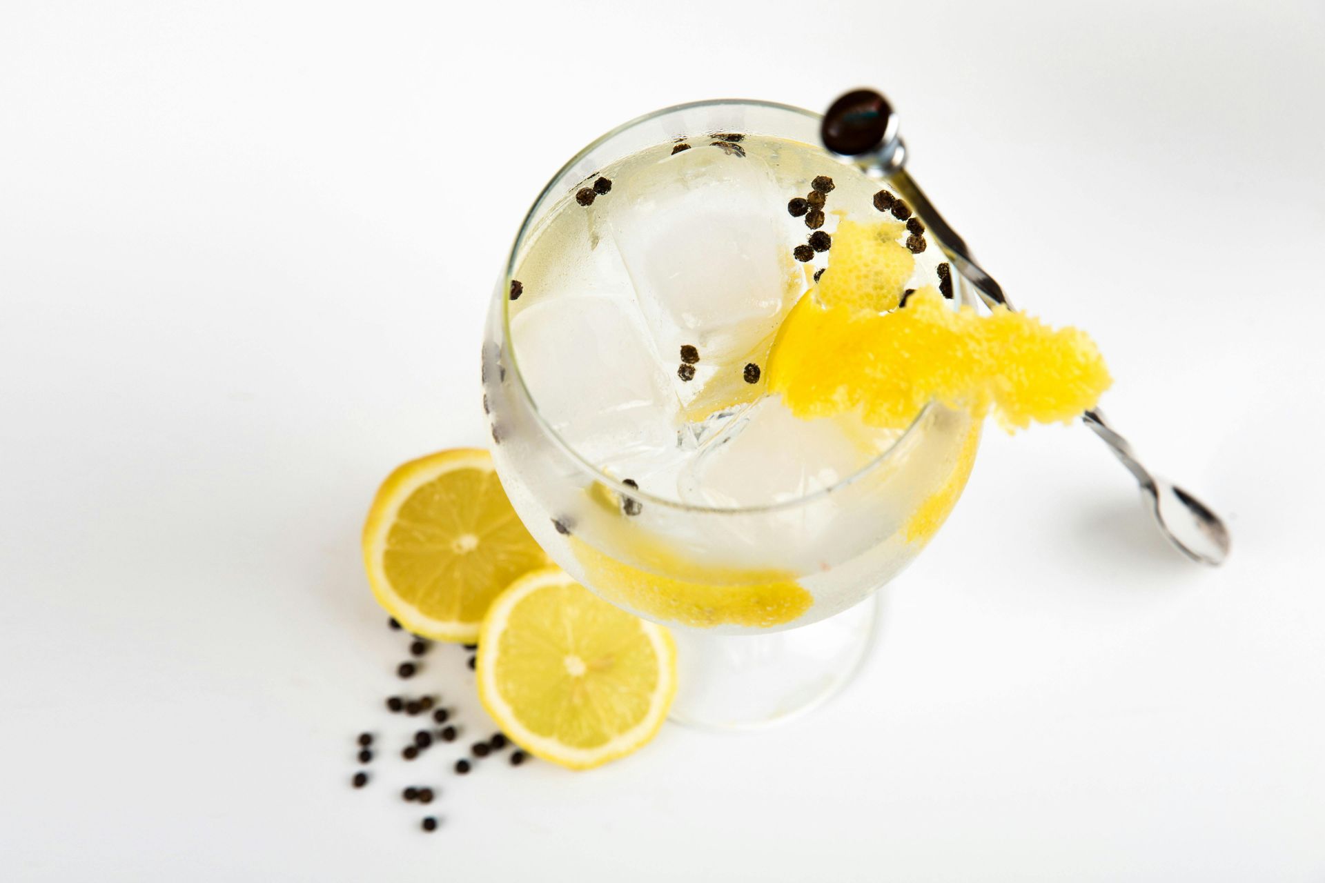 Mula Hotel | Gin and tonic cocktail with lemon garnish and peppercorns, on a white surface.