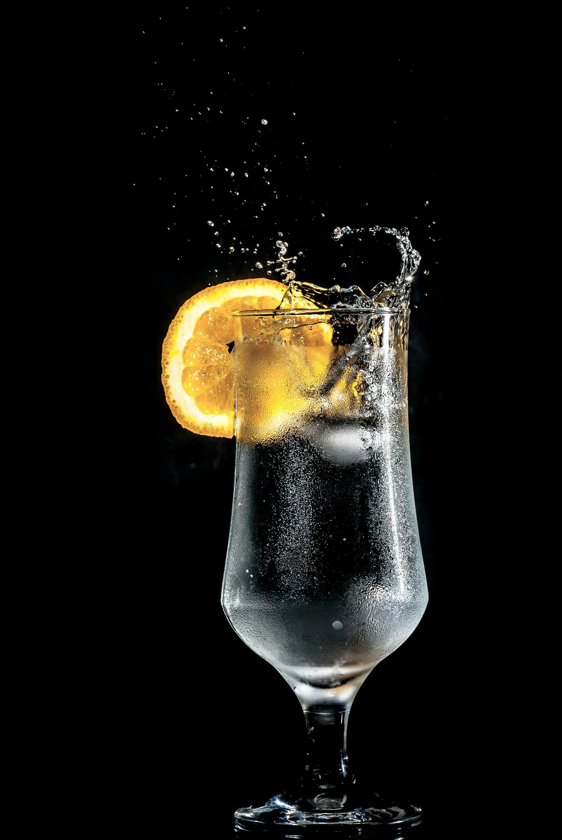 Mula Hotel |Glass of sparkling liquid with lemon slice and ice, splashing against black background.