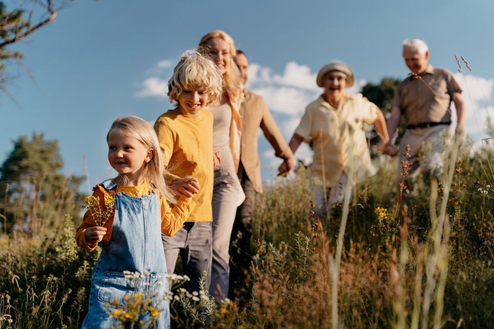Family walking through a grassy field, smiling and carrying flowers in warm sunlight