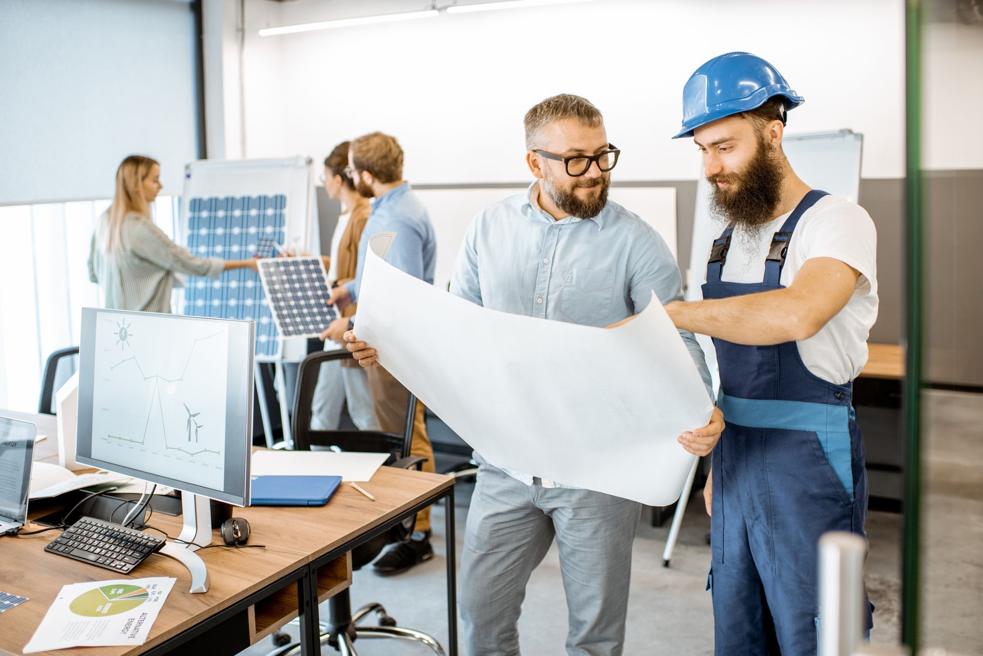 Two engineers in hard hats review a large blueprint in a bright office workspace.