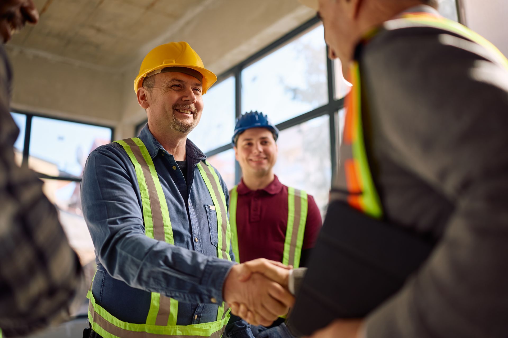Two construction workers shaking hands at a job site, one in a yellow hard hat and safety vest