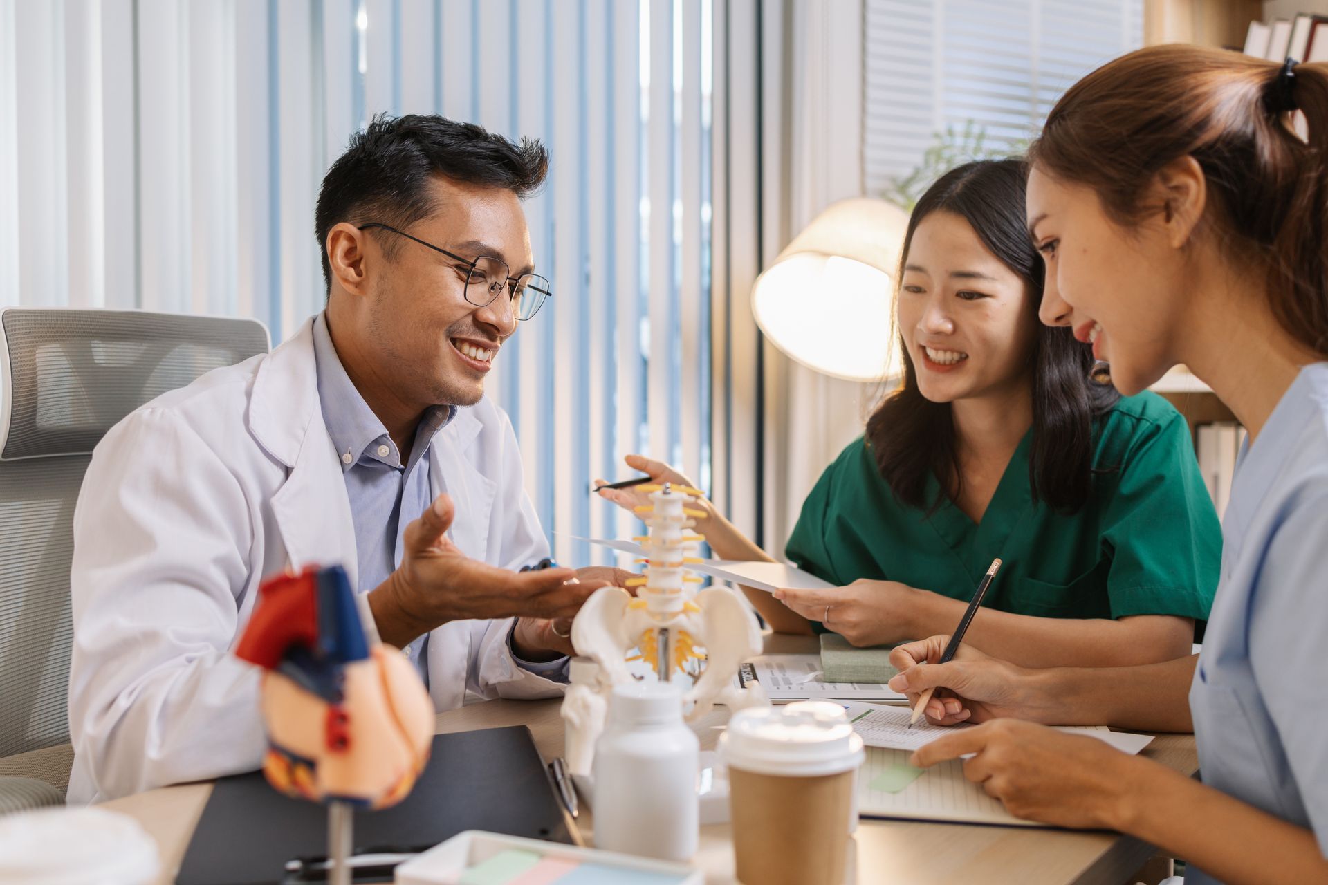 Three people in medical scrubs and a lab coat discuss a dental model at a table with coffee cups.