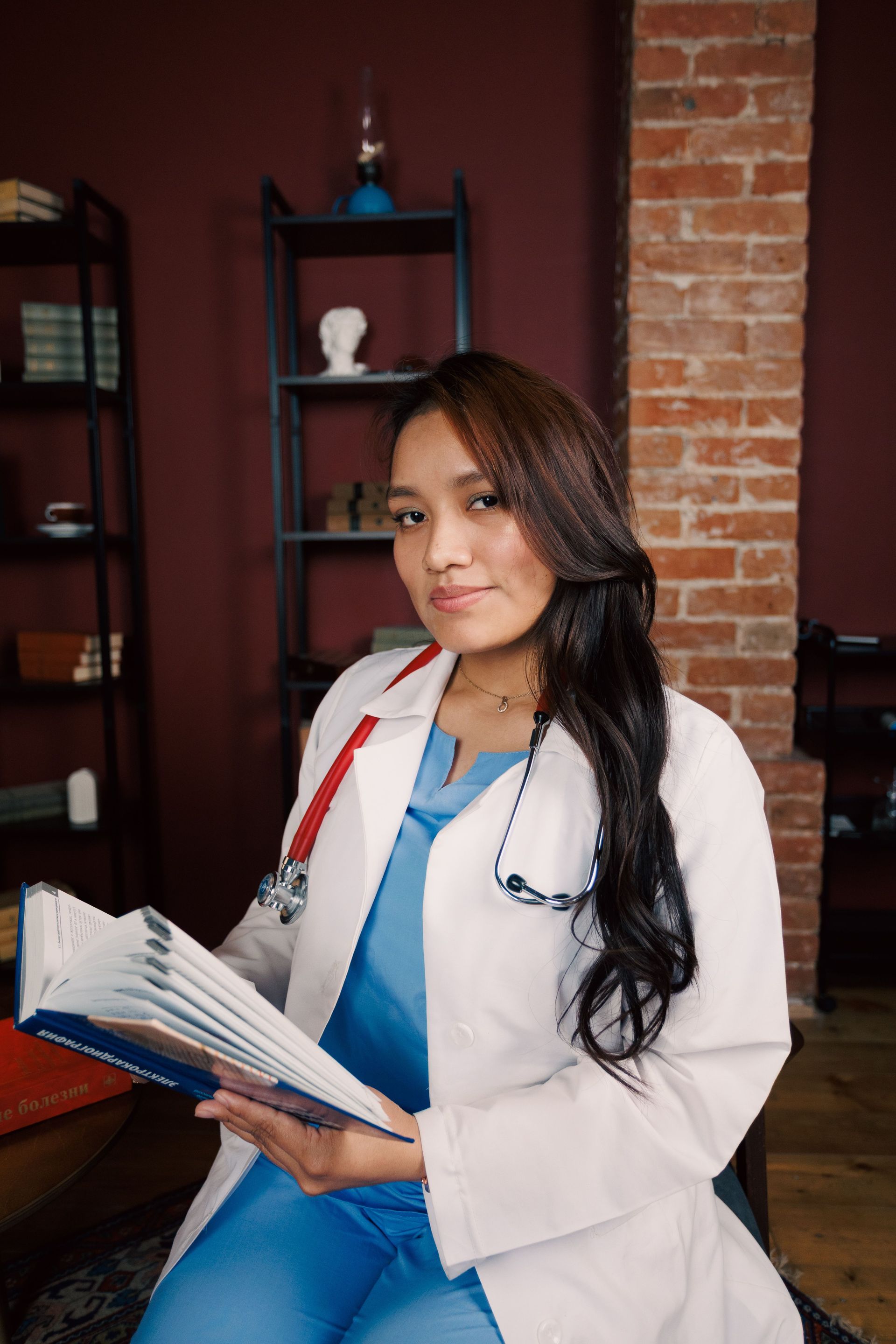 Person in a white coat holding an open book, seated indoors against a brick wall.