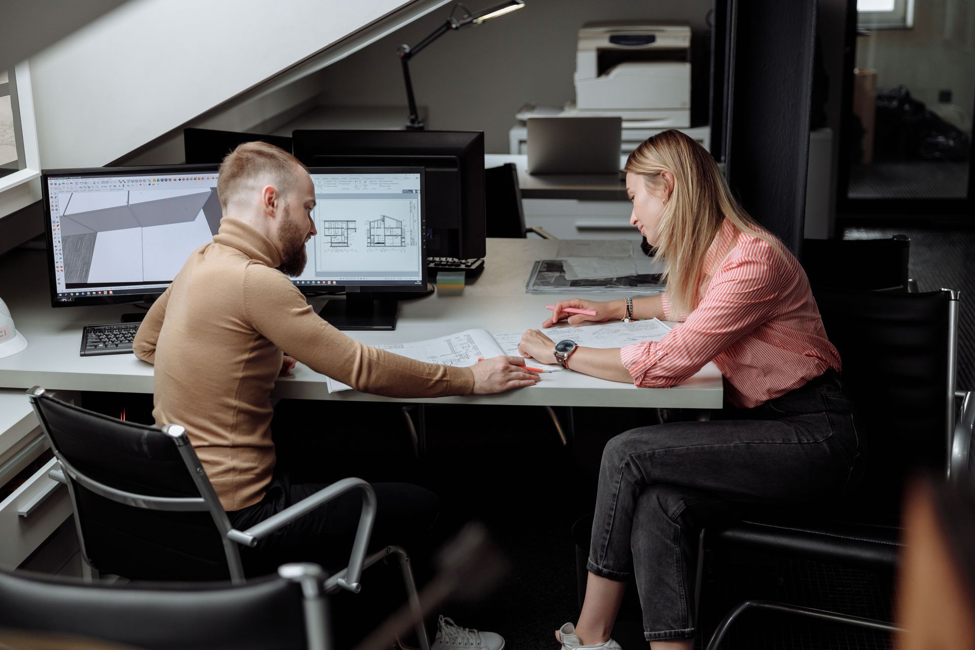 Two people in an office discussing paperwork at a desk with dual monitors