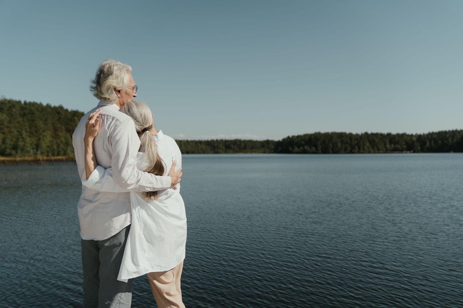 Two people in white clothing embrace beside a calm lake under a clear sky