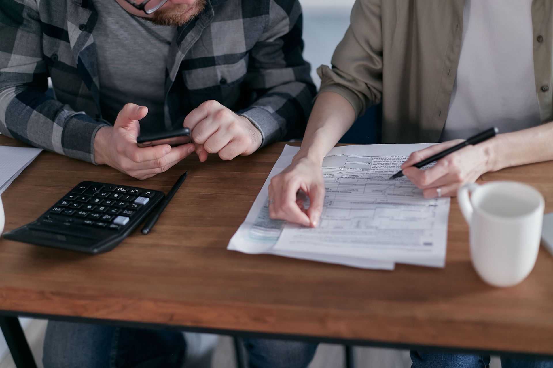Two people at a table reviewing paperwork, one using a phone, with a calculator and coffee mug nearby.