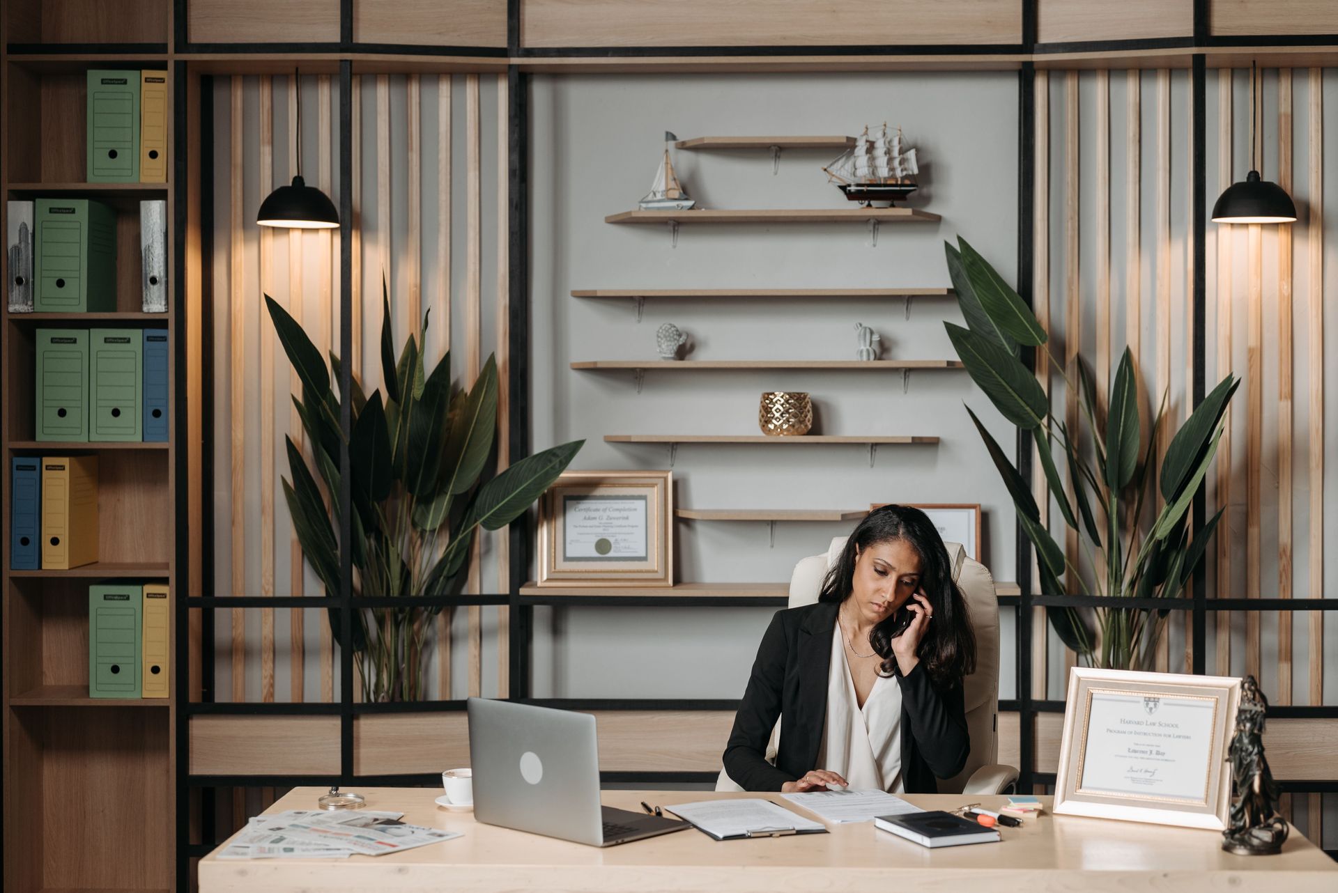 Woman working at a desk in a modern office with plants, shelves, and a laptop