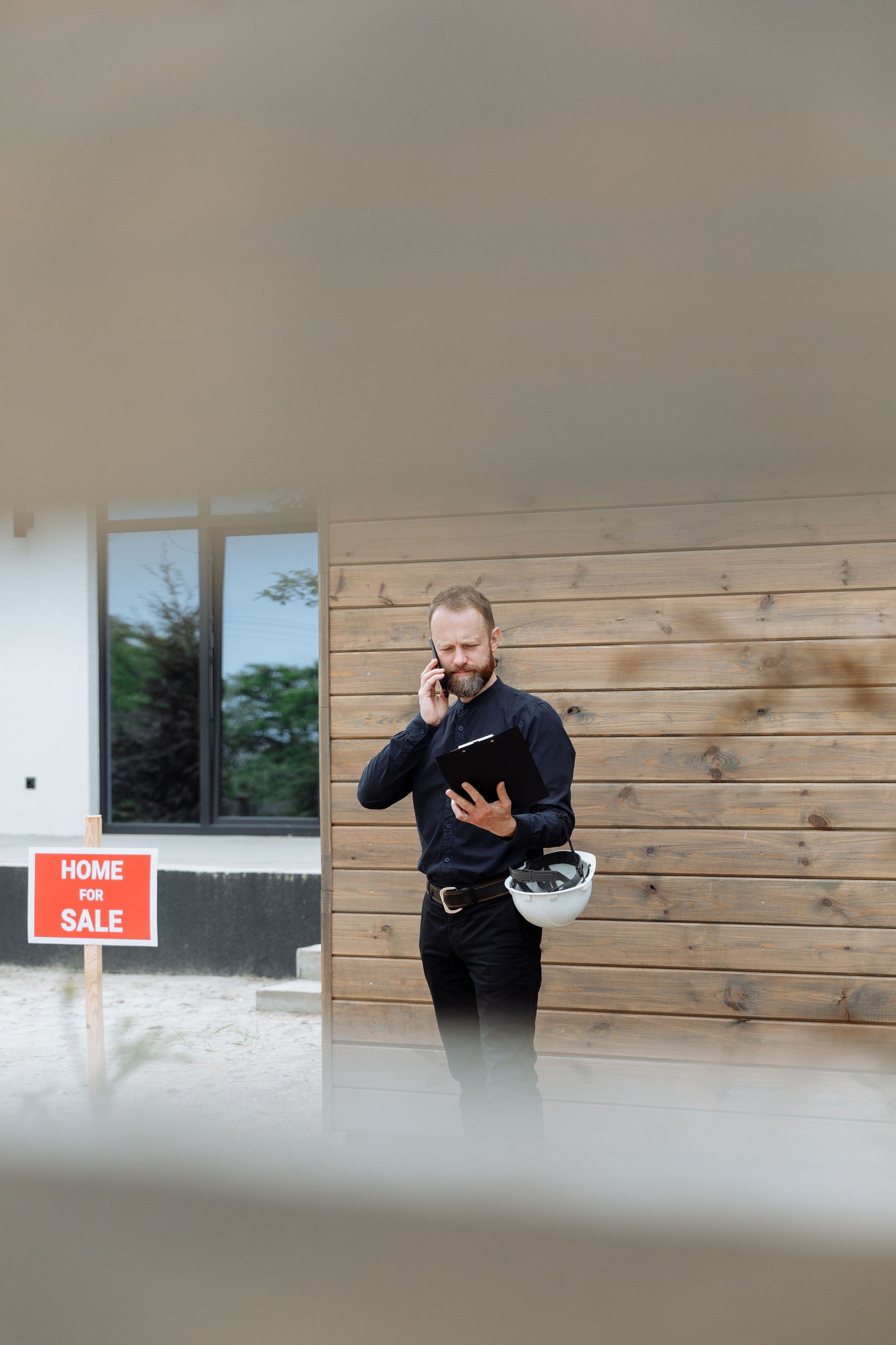 Person standing outside a building, talking on a phone and holding a helmet, with a “For Sale” sign nearby.