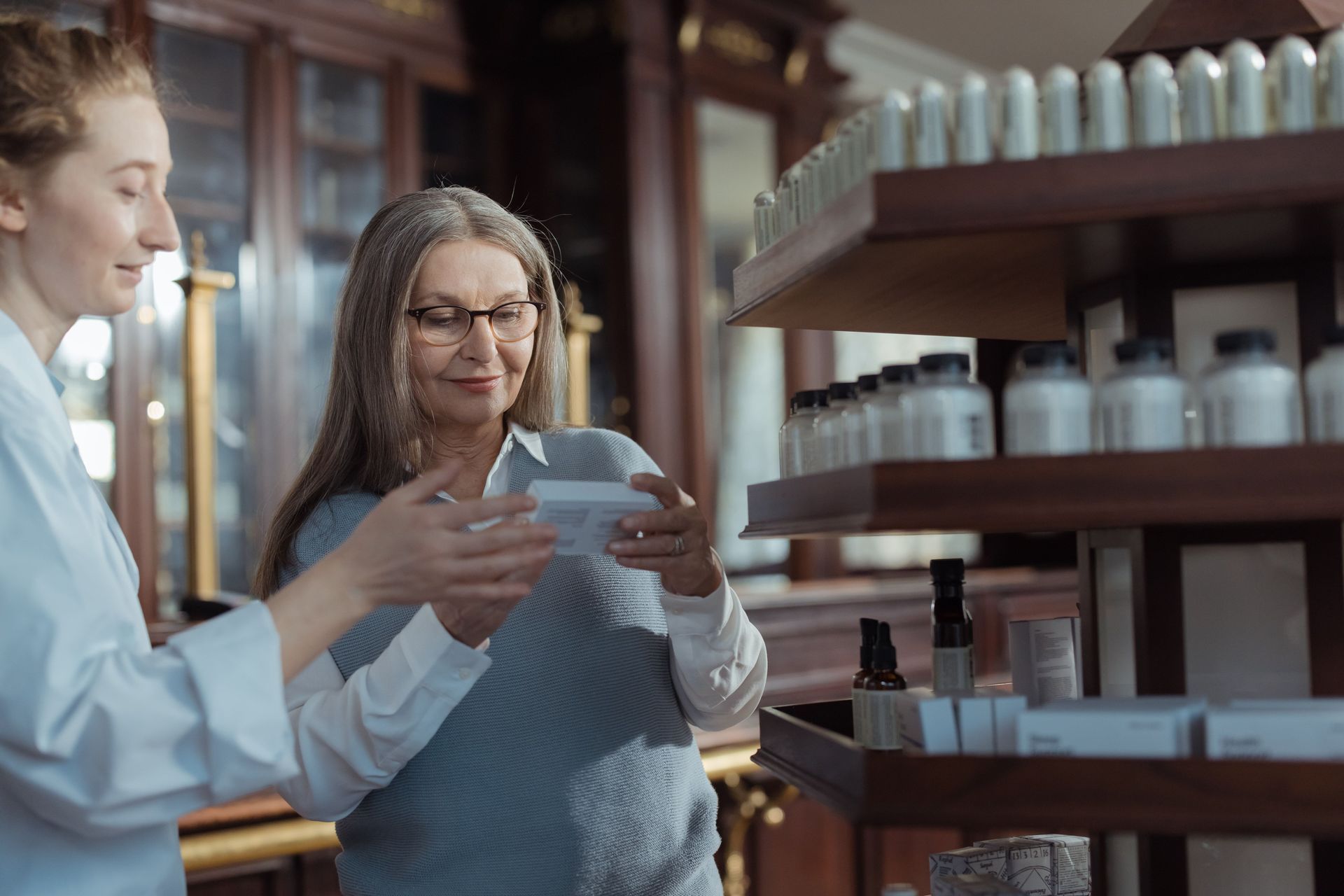 Two lab workers examine a small vial in a pharmacy aisle.