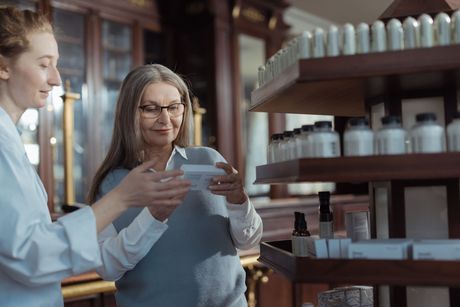 Two lab workers examine a small vial in a pharmacy aisle.