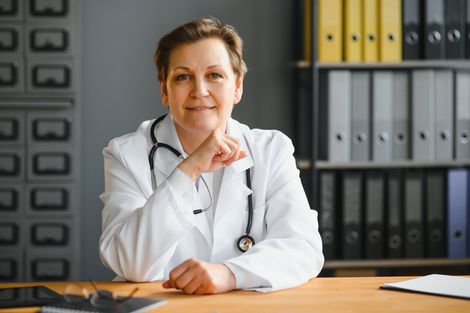 Doctor in a white coat seated at a desk in an office, with folders on shelves behind her.