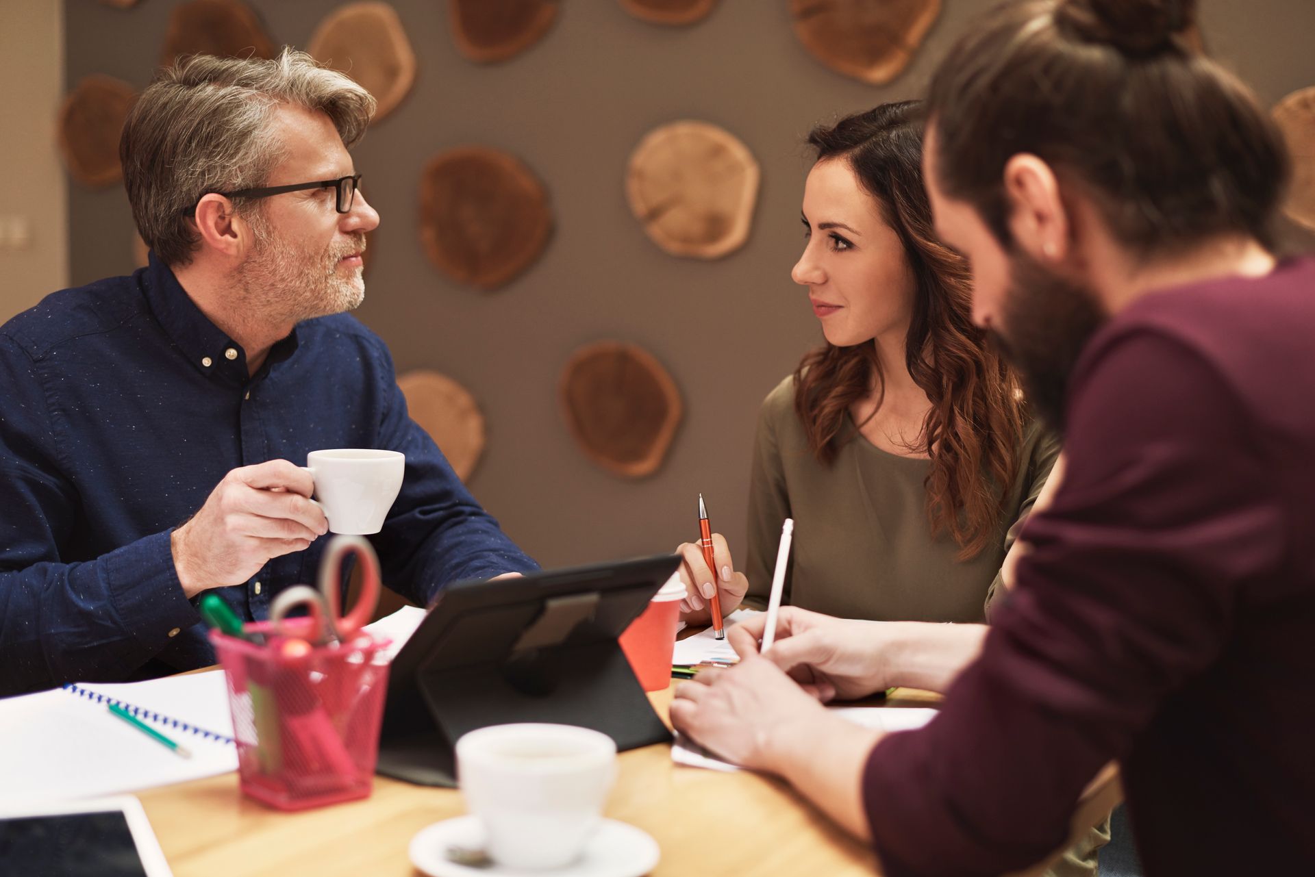 Three people talking over coffee at a table with notebooks and a tablet in a warm meeting room