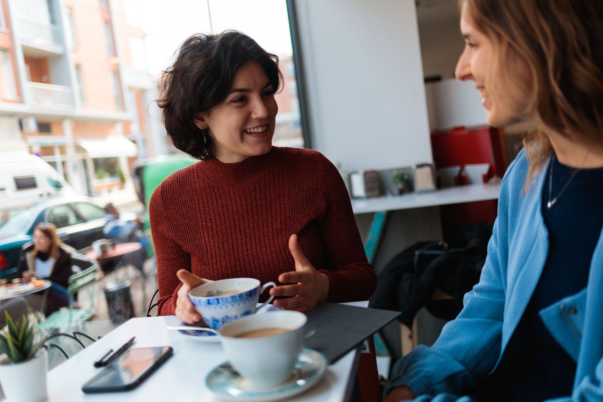 Two people chatting at a café table, one holding a cup and smiling, with coffee cups and a phone on the table