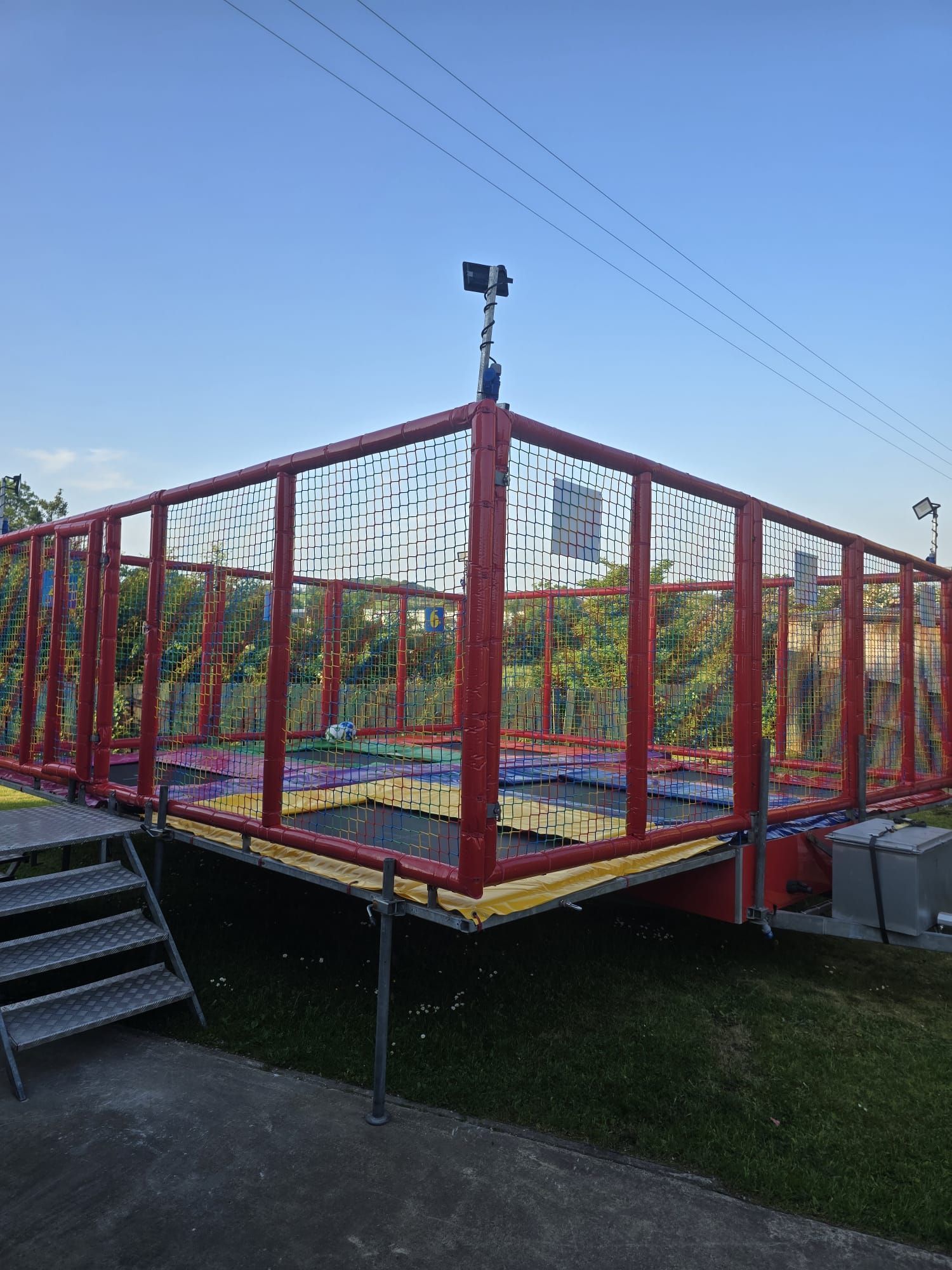 Red metal platform stage with netting, lights on a pole, and steps beside it outdoors on grass