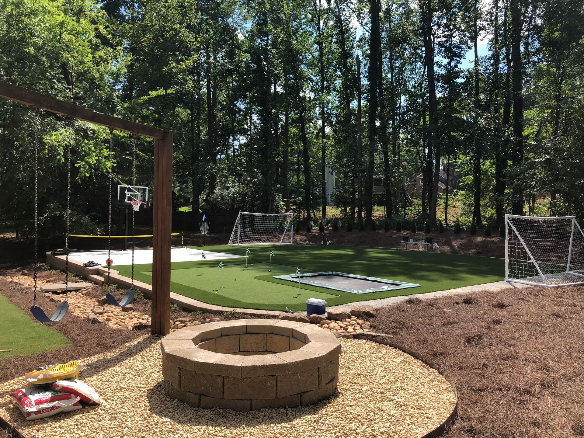 Backyard with artificial turf, trampoline, soccer goals, and a stone fire pit surrounded by mulch and trees