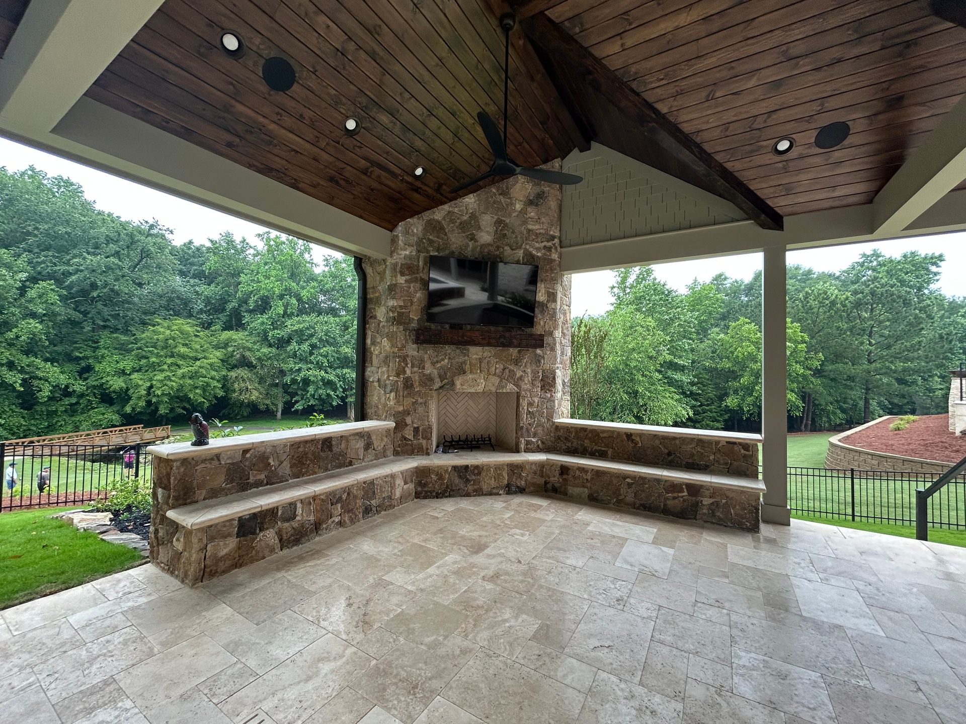 Covered outdoor pavilion with stone seating, a fireplace, and trees beyond the open sides
