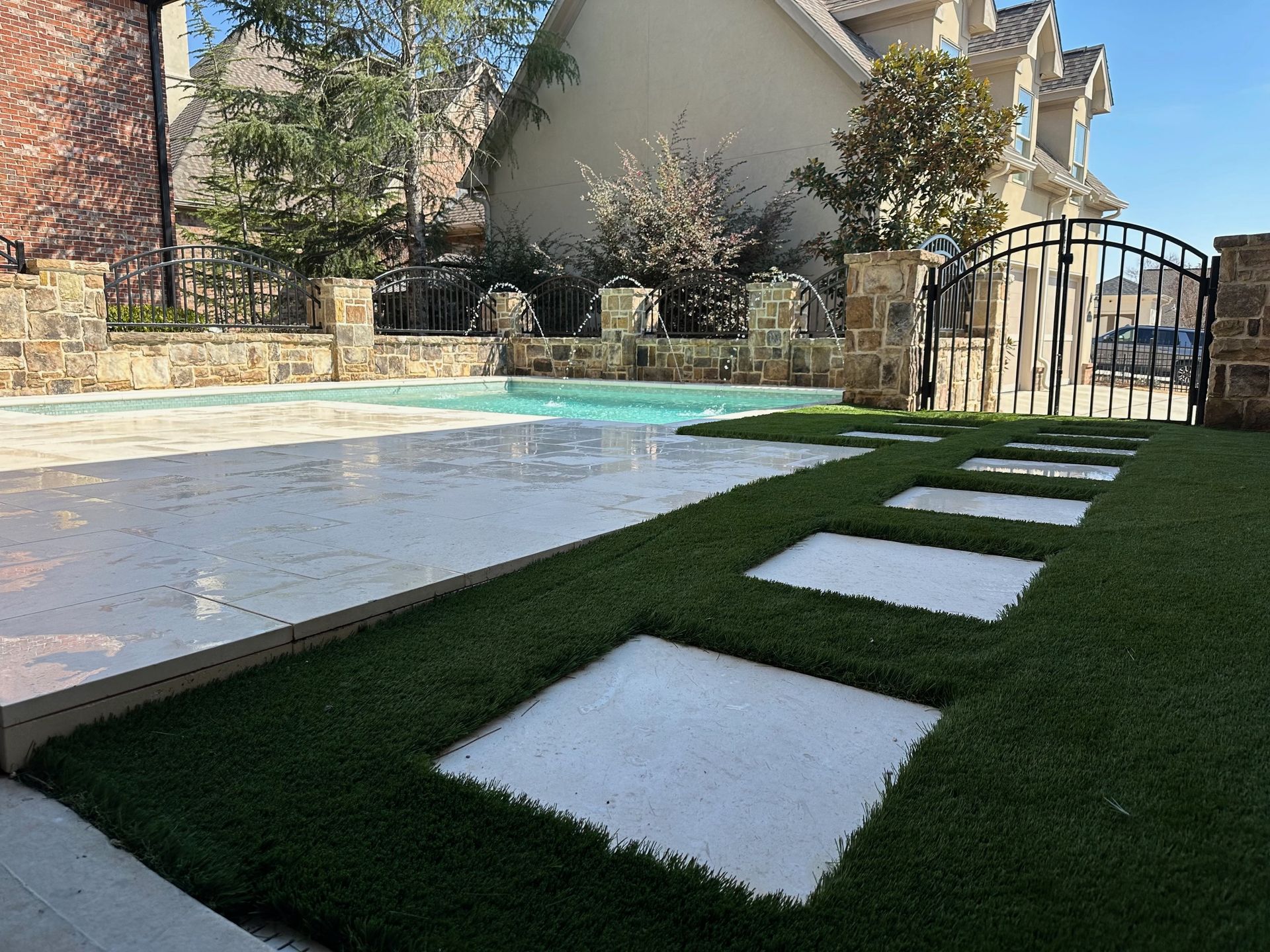 Backyard pool with white stepping stones in green artificial grass, stone patio, and wrought-iron gate