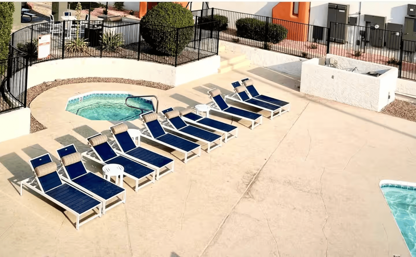 Empty pool deck with blue lounge chairs beside a small hot tub and black fencing.