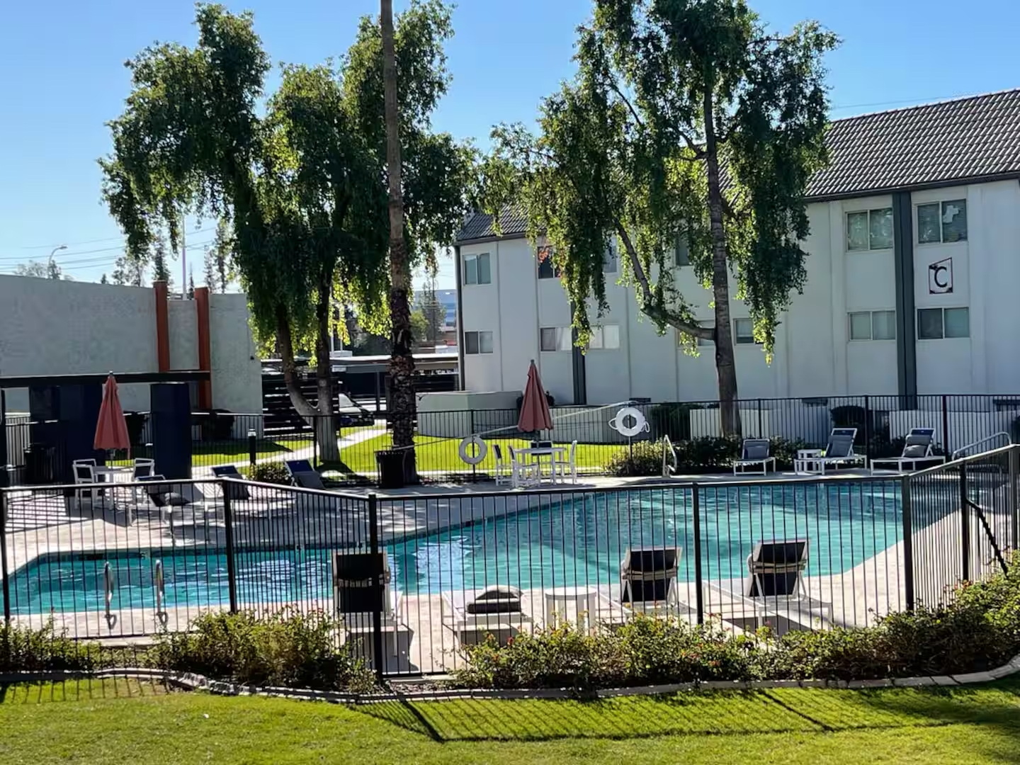 Outdoor swimming pool with lounge chairs, umbrellas, and trees beside apartment buildings.