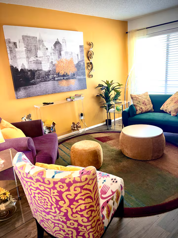 Colorful living room with patterned chairs, round ottomans, and wall art on a mustard-yellow wall.