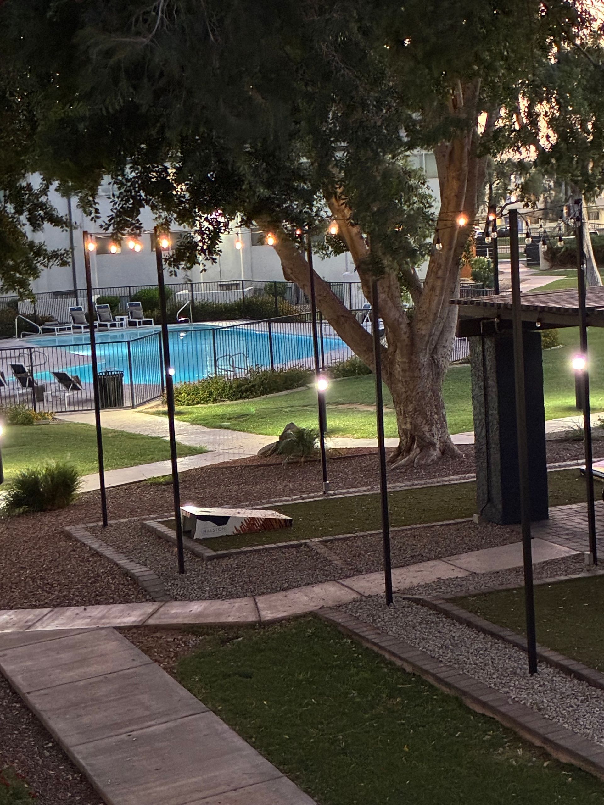 Lit outdoor pool beside trees and walkways at dusk, with lamp posts and a patio area