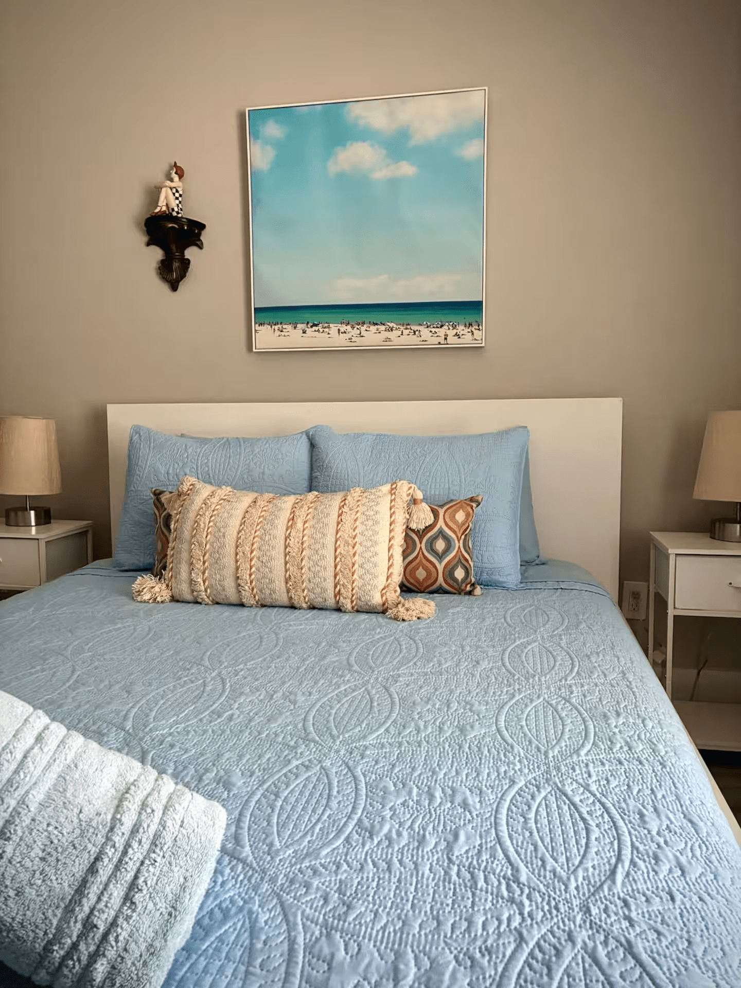 Cozy bedroom with a blue bedspread, patterned pillows, and a beach painting above the headboard
