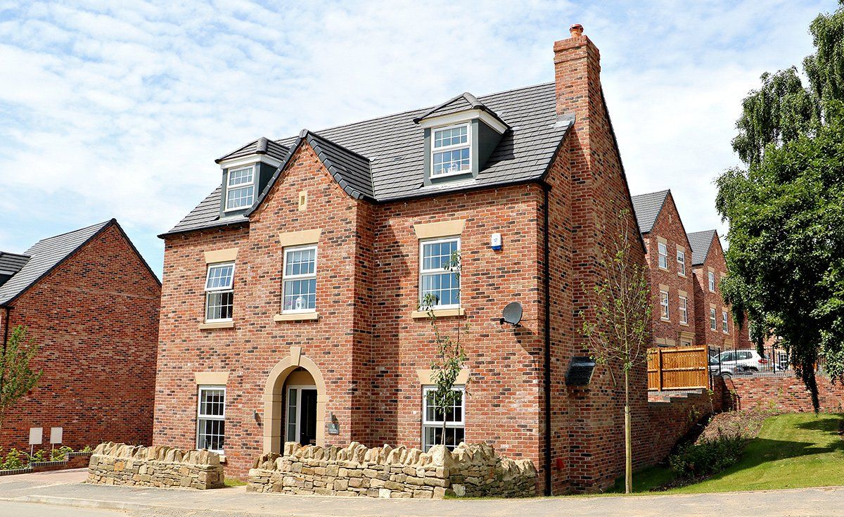 Brick house with dormers, a chimney, and a stone wall in front, under a blue sky.