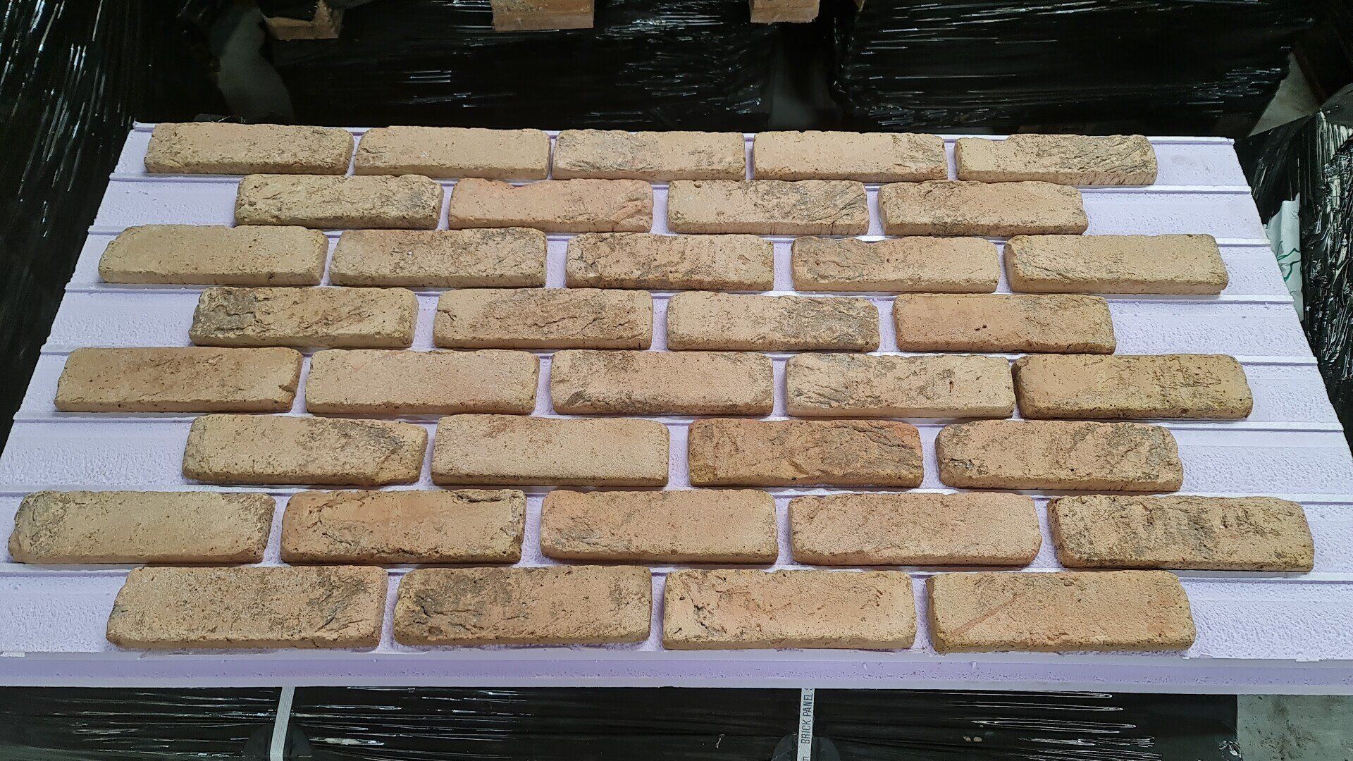 Brick tiles in a running bond pattern, beige with dark specks, displayed on a white foam board.