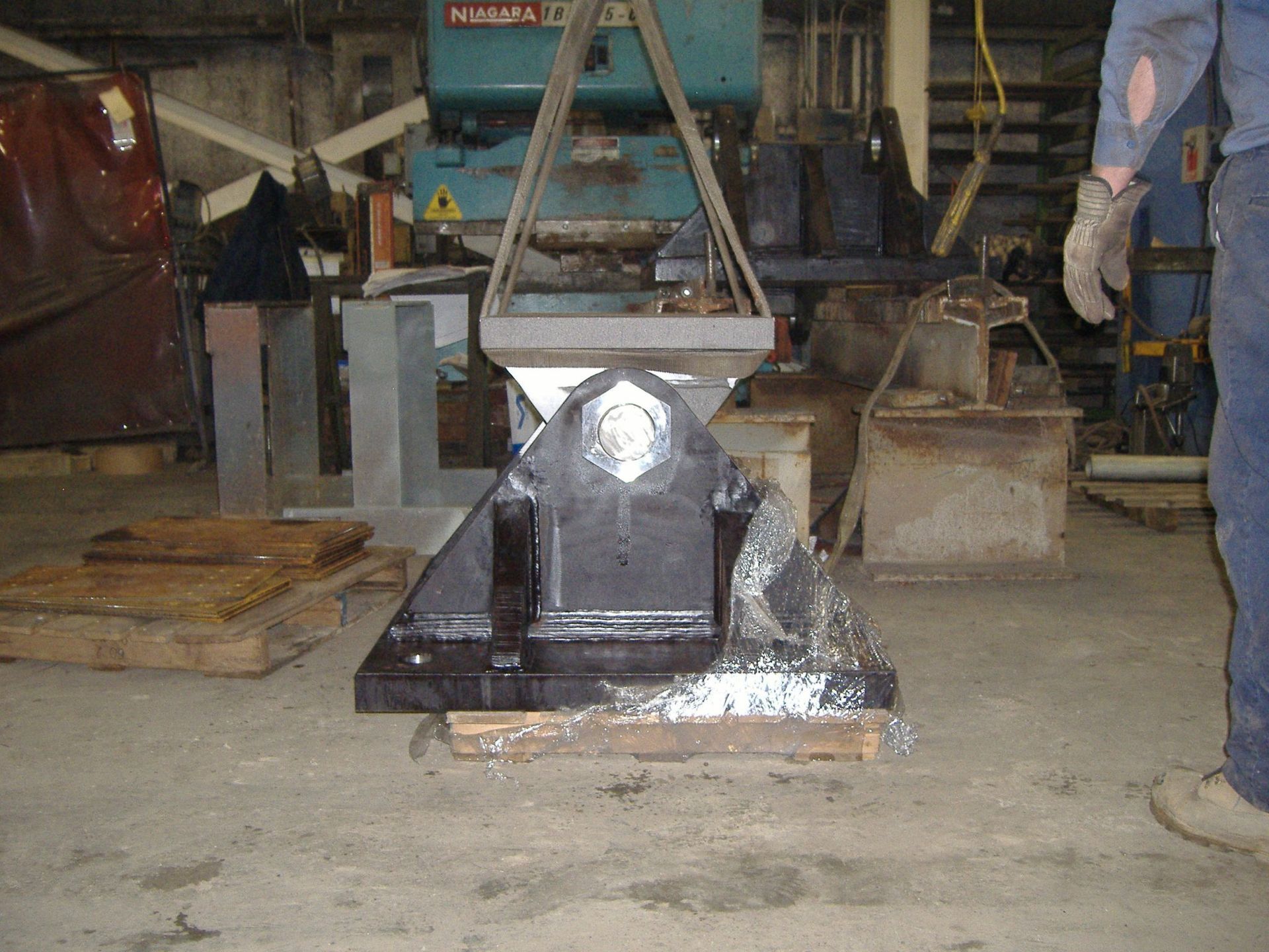 A large black metal part suspended by a crane in a workshop. A worker is partially visible on the right.