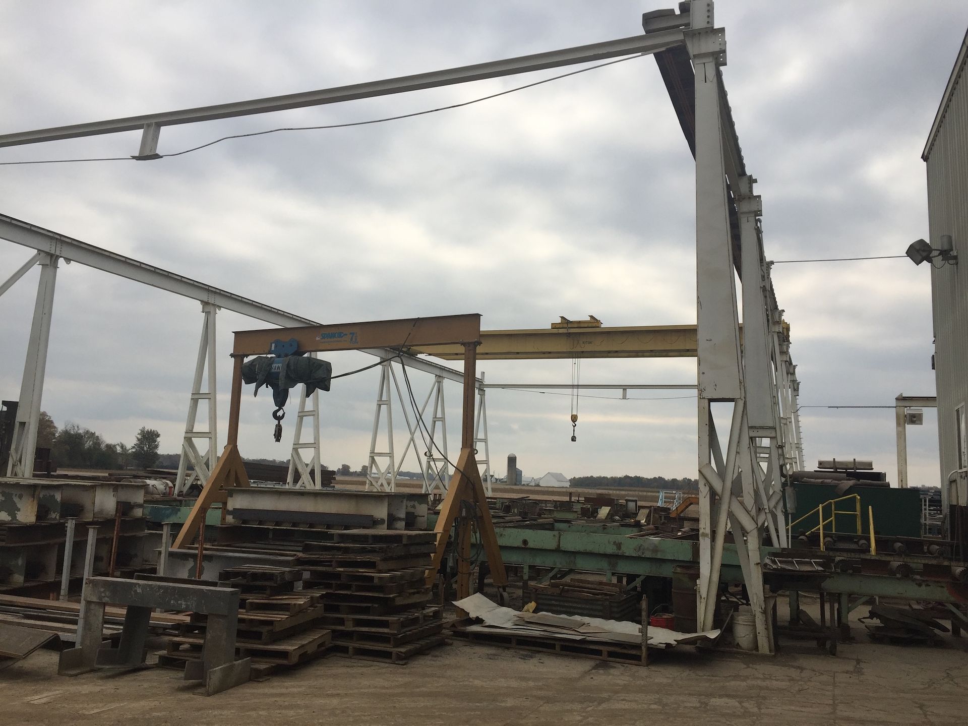Industrial yard with overhead cranes, machinery, and material stacks under a cloudy sky.