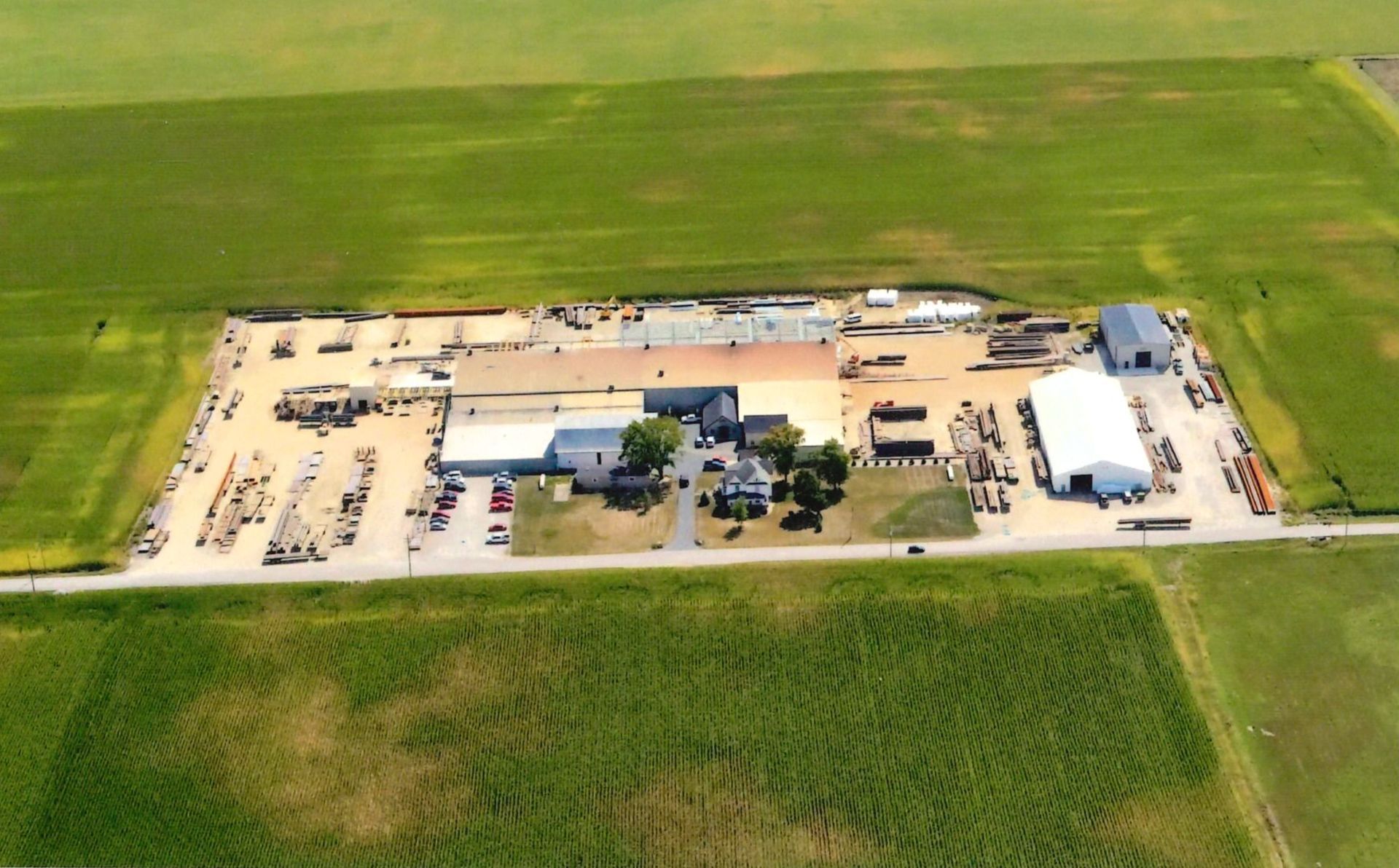 Aerial view of a farm or industrial complex with buildings, gravel lot, vehicles, and surrounding green fields.