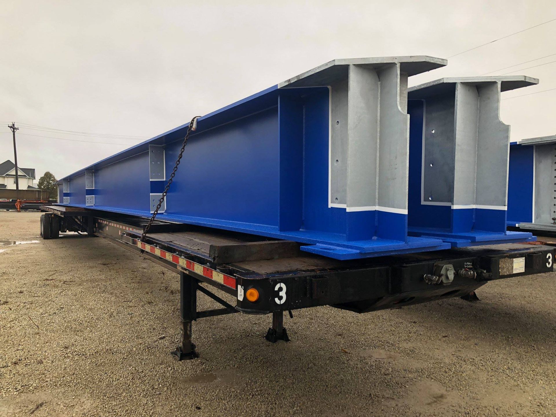 Blue and gray steel beams on a flatbed trailer, parked on gravel.