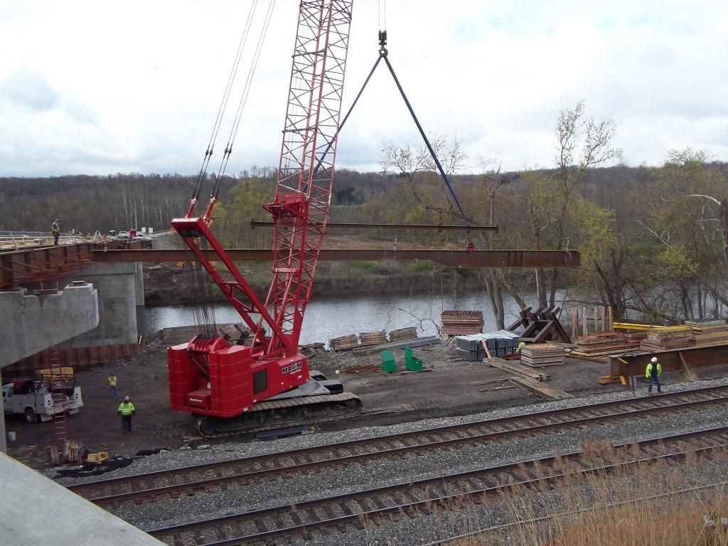 Red crane lifting a steel beam during bridge construction over a river. Construction workers nearby.