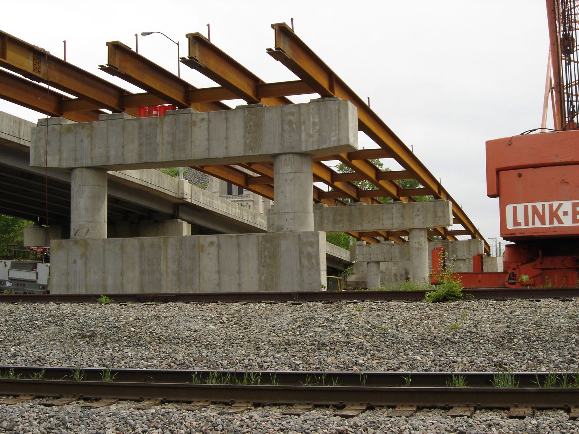 Bridge construction over railroad tracks, with concrete supports and steel beams, near an orange crane.
