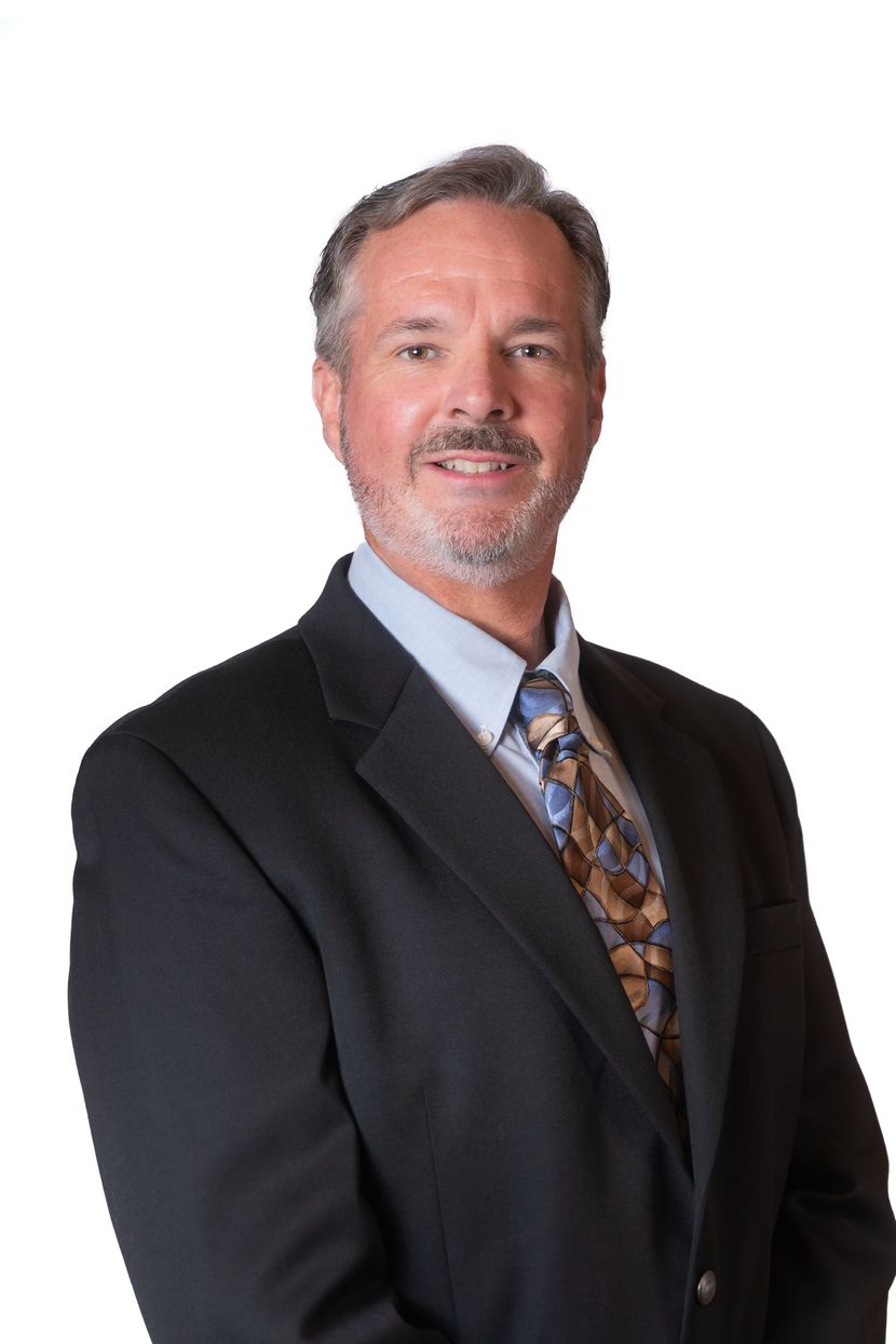 Man in a black suit and patterned tie, smiling against a plain white background