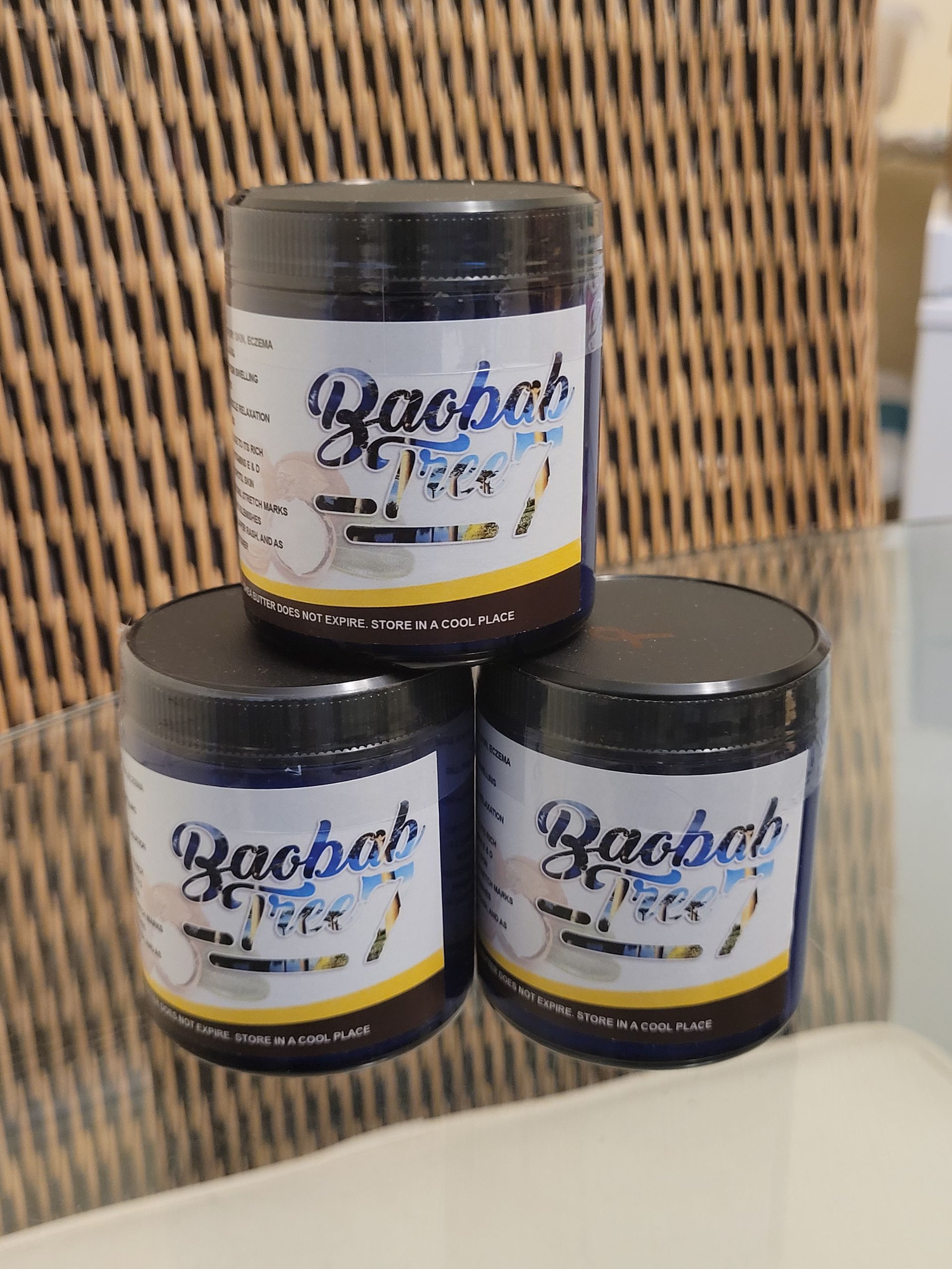 Three black-lidded cosmetic jars stacked on a wicker background, with white and gold labels.